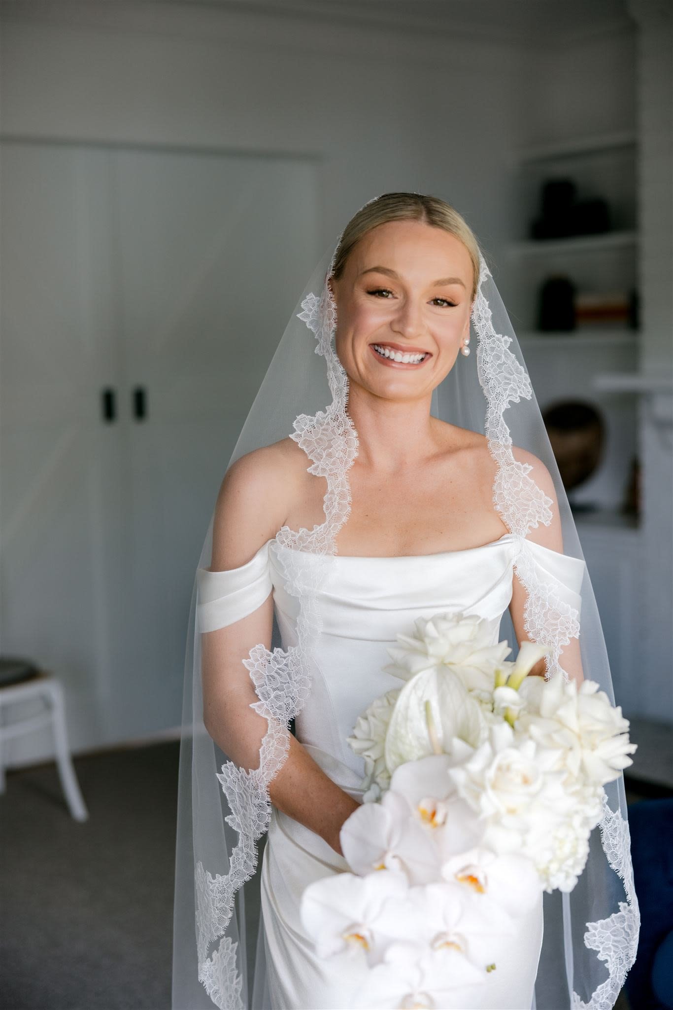 Bride in a white off-the-shoulder gown with lace-edged veil, holding a bouquet of white flowers, smiling indoors.