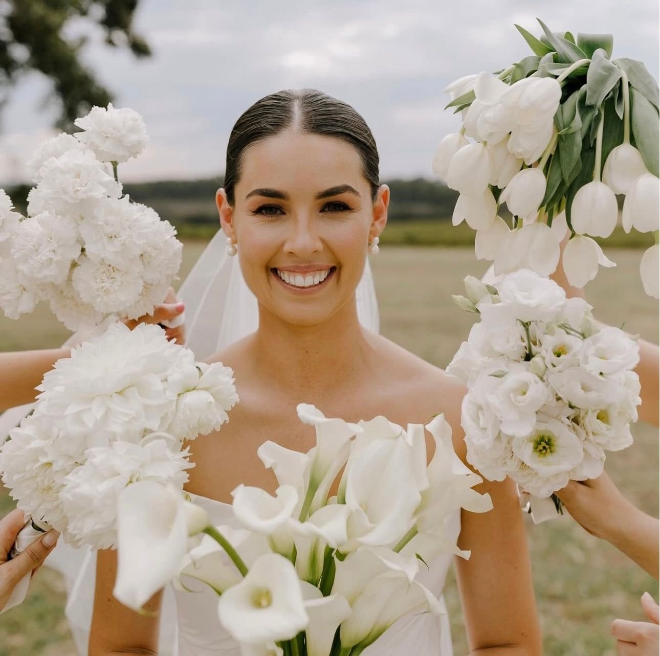 Bride surrounded by white flowers, including tulips, calla lilies, and roses, smiling outdoors.