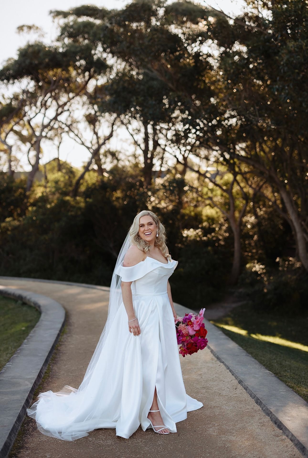 Bride in white gown holding colorful bouquet on outdoor path with trees in background.
