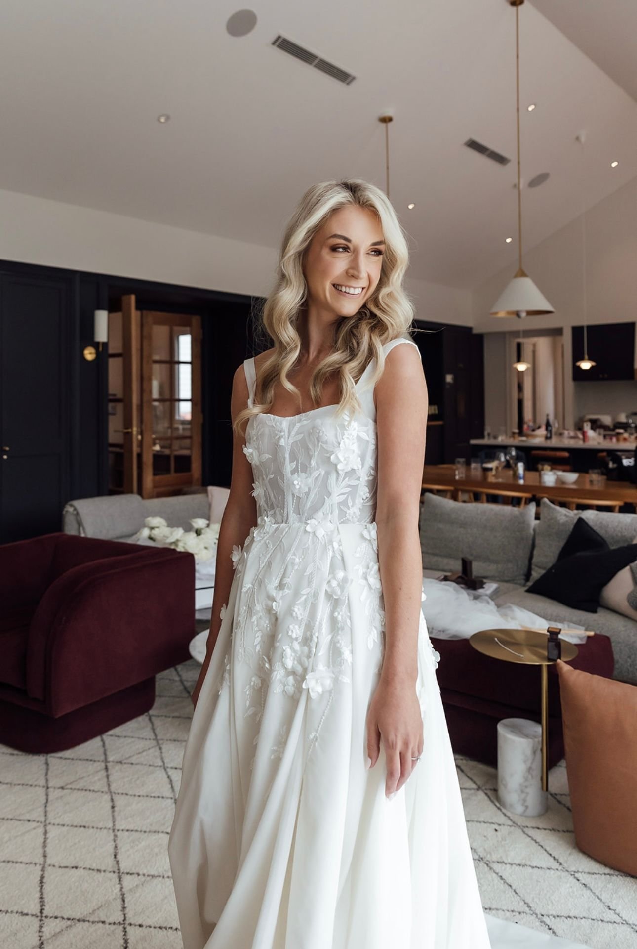 A bride in a white floral wedding dress posing in a modern living room with dark furniture and pendant lights.