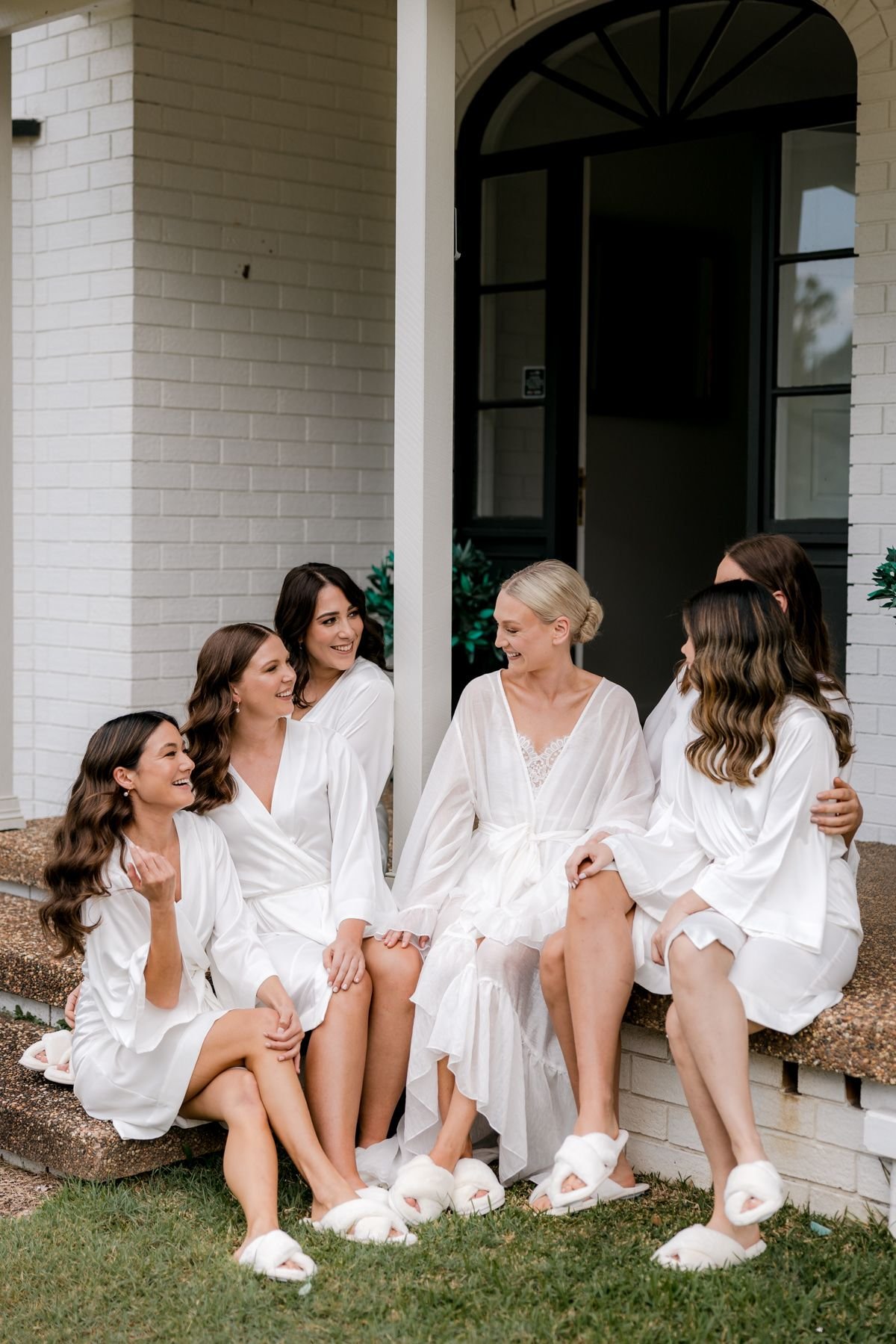 A group of six women in white robes and slippers sitting on steps, smiling and talking, outside a brick building.
