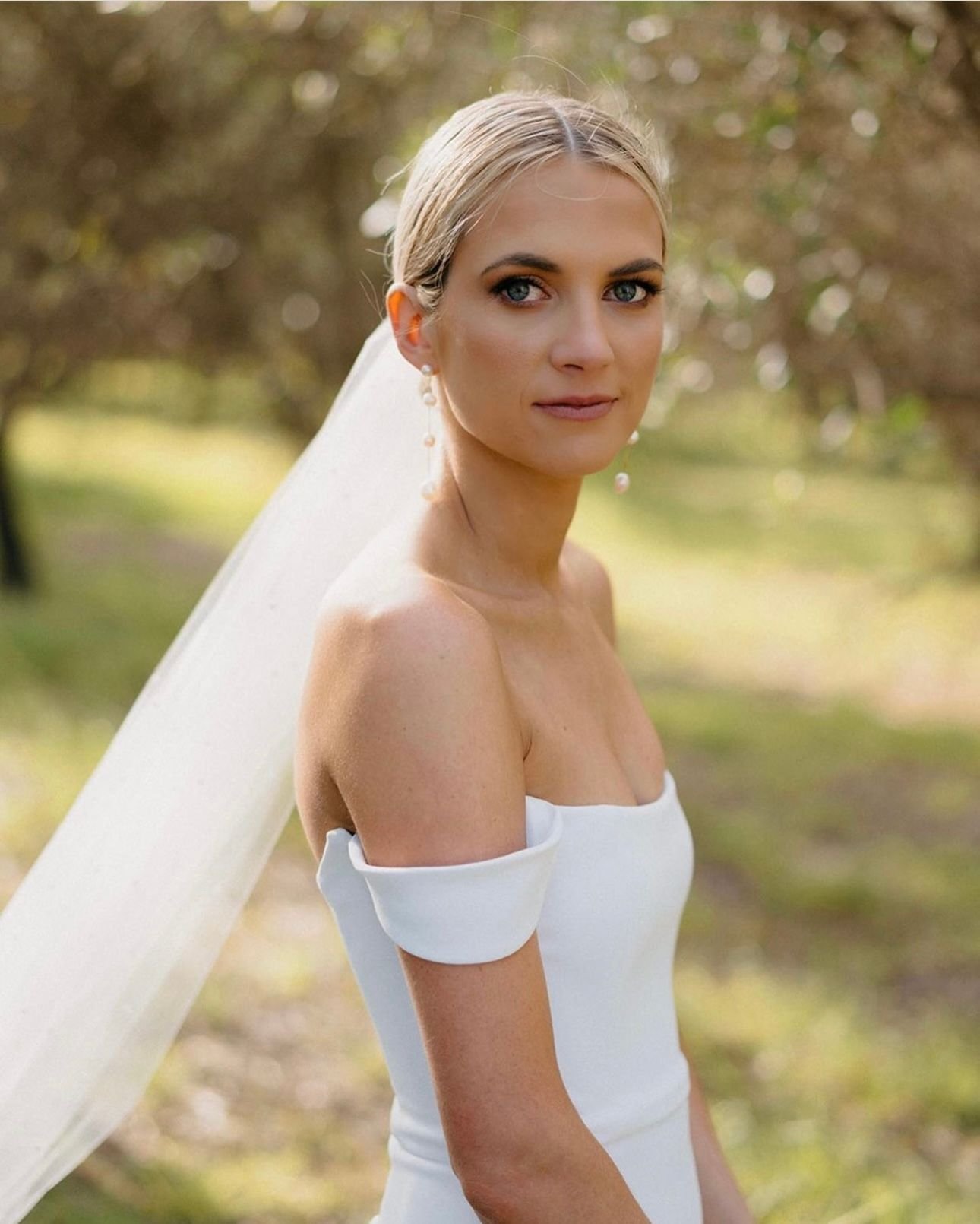 Bride in white off-shoulder wedding dress with veil, outdoor setting.