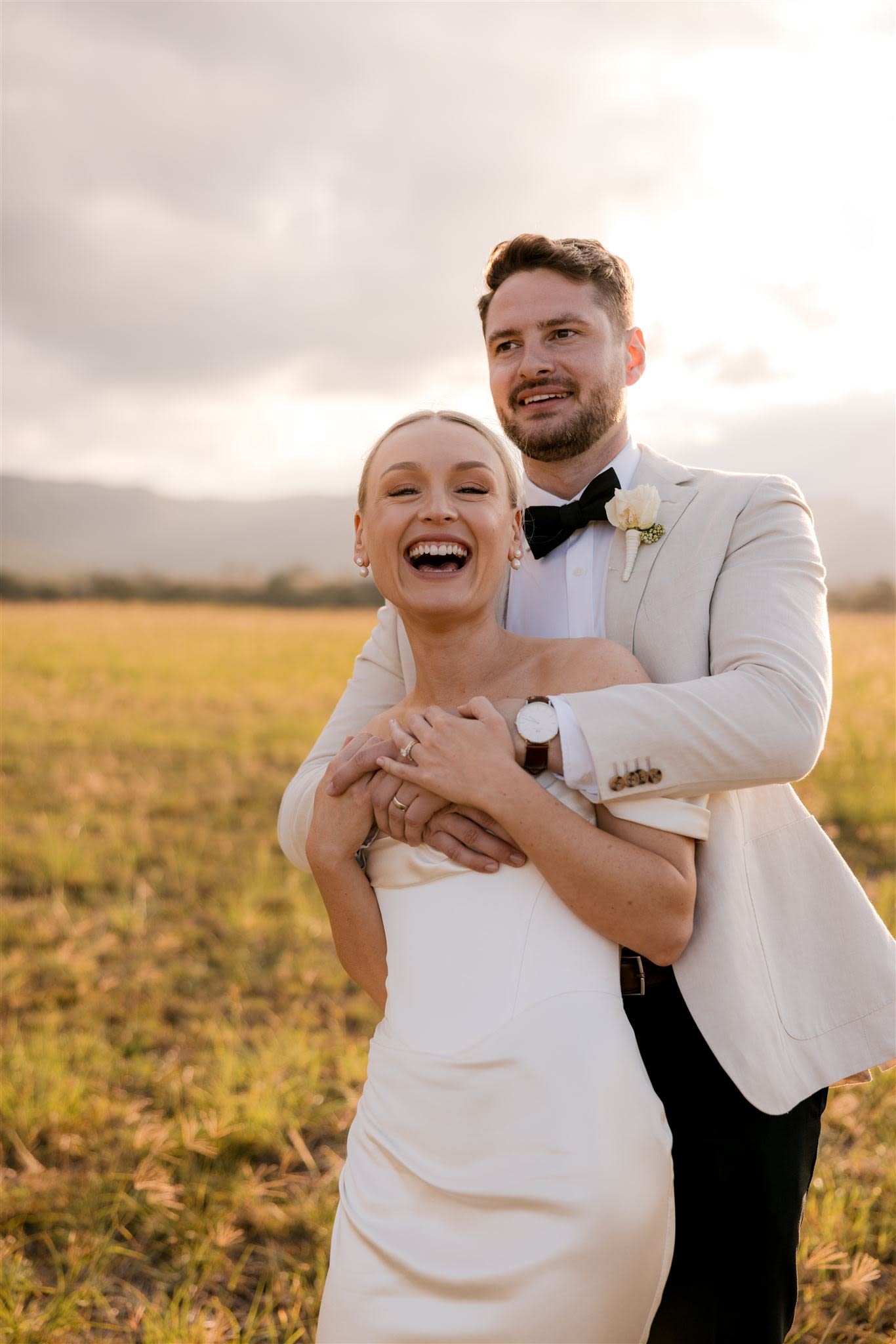 Smiling couple in wedding attire embracing outdoors in a field, with the sun setting behind them.
