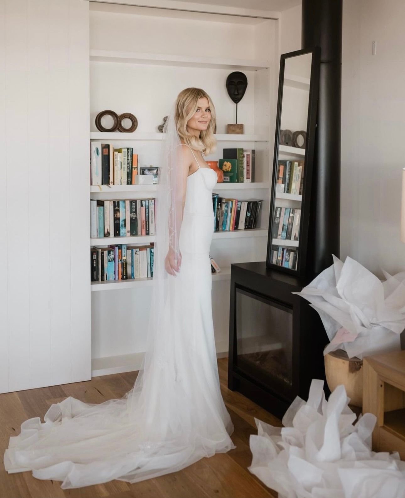 A woman in a white wedding dress and veil stands in front of a bookshelf and a tall mirror. The background includes a wood floor, books, and decorative items on shelves. Crumpled white paper is scattered on the floor near a fireplace.