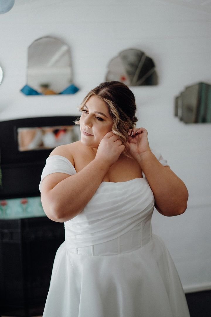Bride in white gown adjusting earring, with styled hair in a room with mirrors.