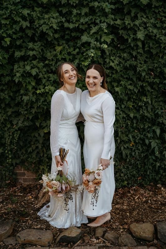 Two women in white dresses, holding bouquets, standing in front of a leafy wall.