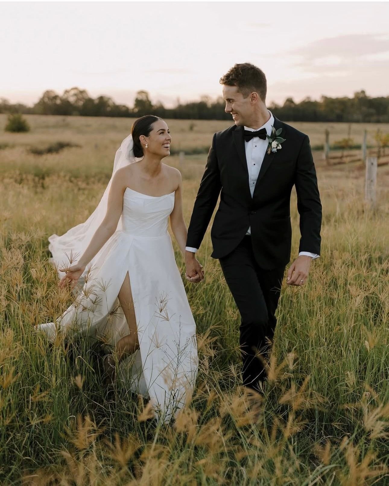 Bride and groom in wedding attire walking through a grassy field, holding hands, and smiling at each other.