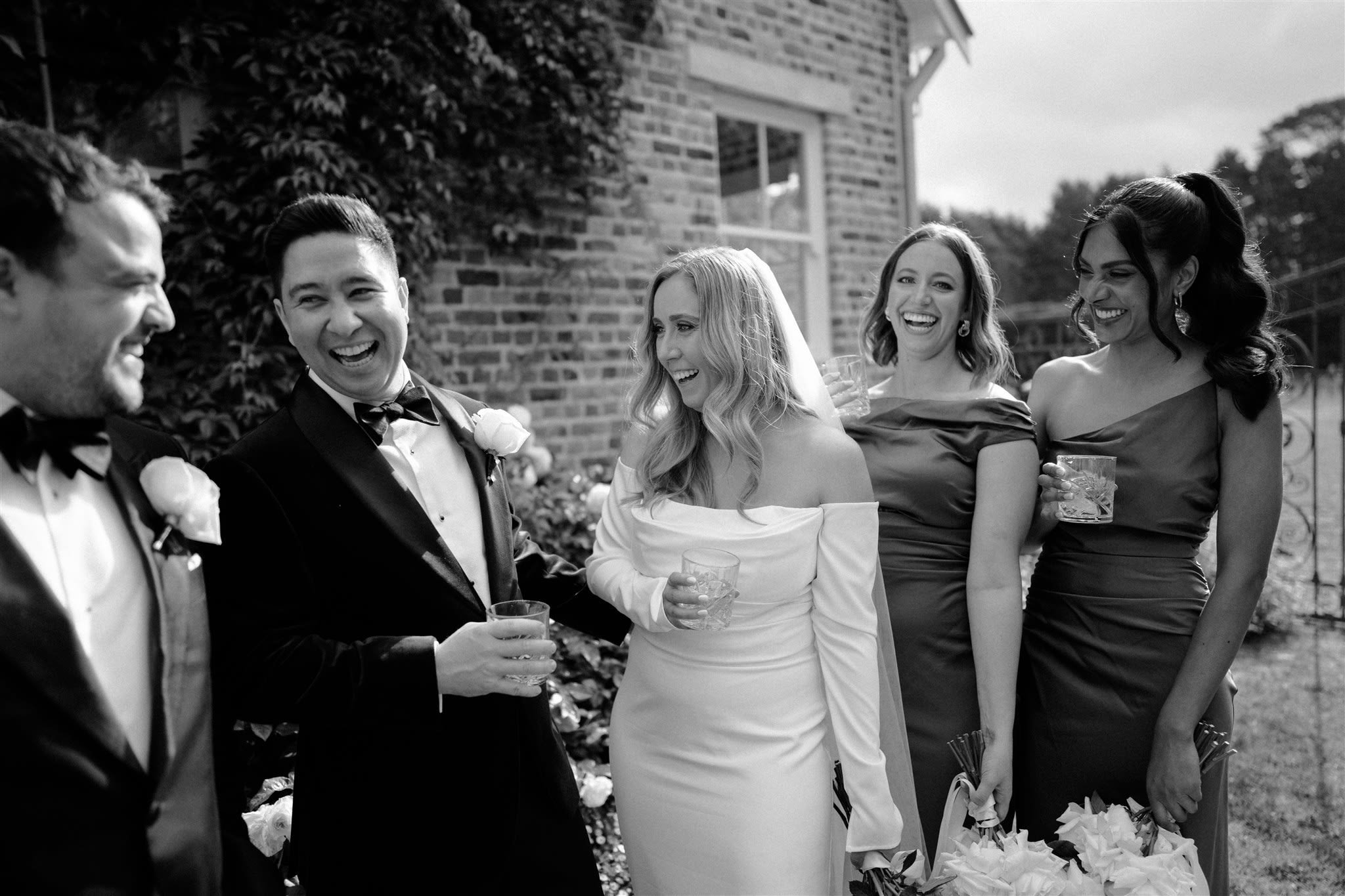 Black and white photo of a bride and wedding party laughing outdoors, with the bride in a white dress and veil, and others in formal attire holding drinks.