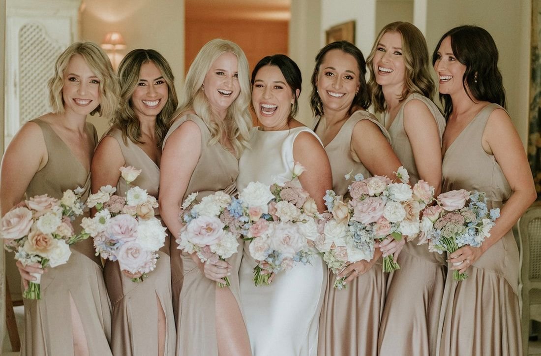 Bride with bridesmaids in matching dresses holding bouquets, smiling indoors.