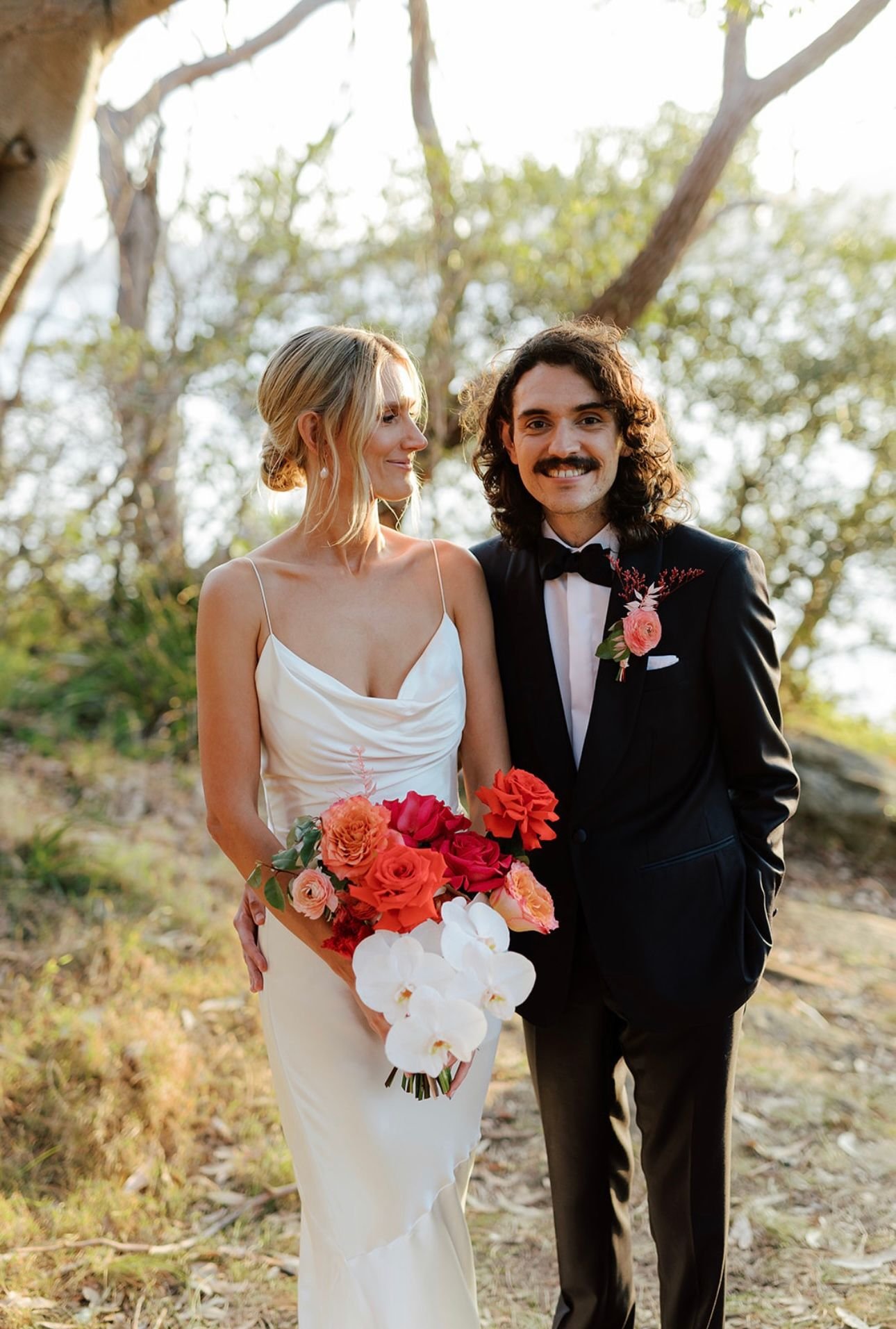 Bride and groom standing together in a garden, bride holding a bouquet of colorful flowers, wearing a white dress and groom in a black suit.