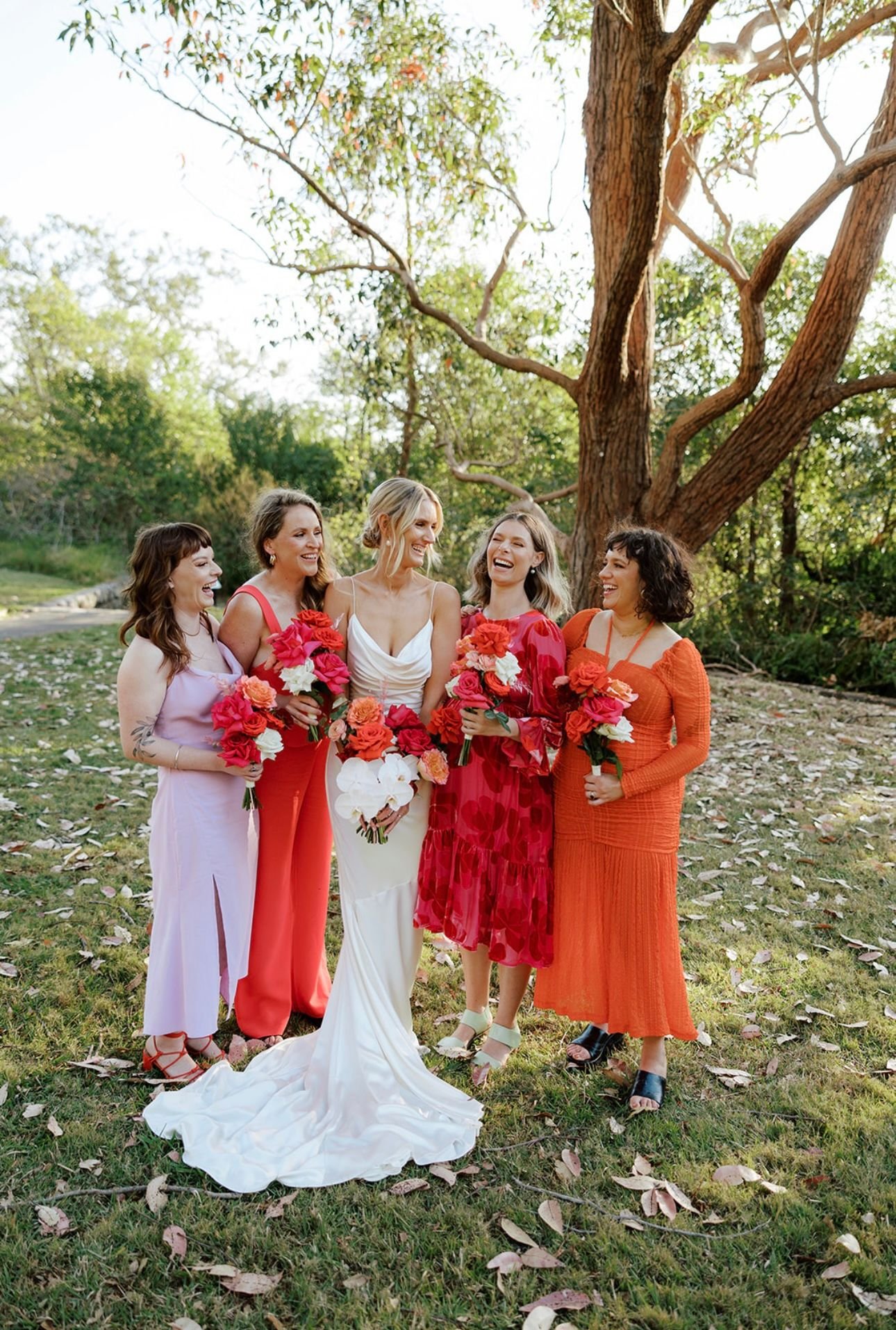 Bride with bridal party holding bouquets, standing outdoors on grass, surrounded by trees.