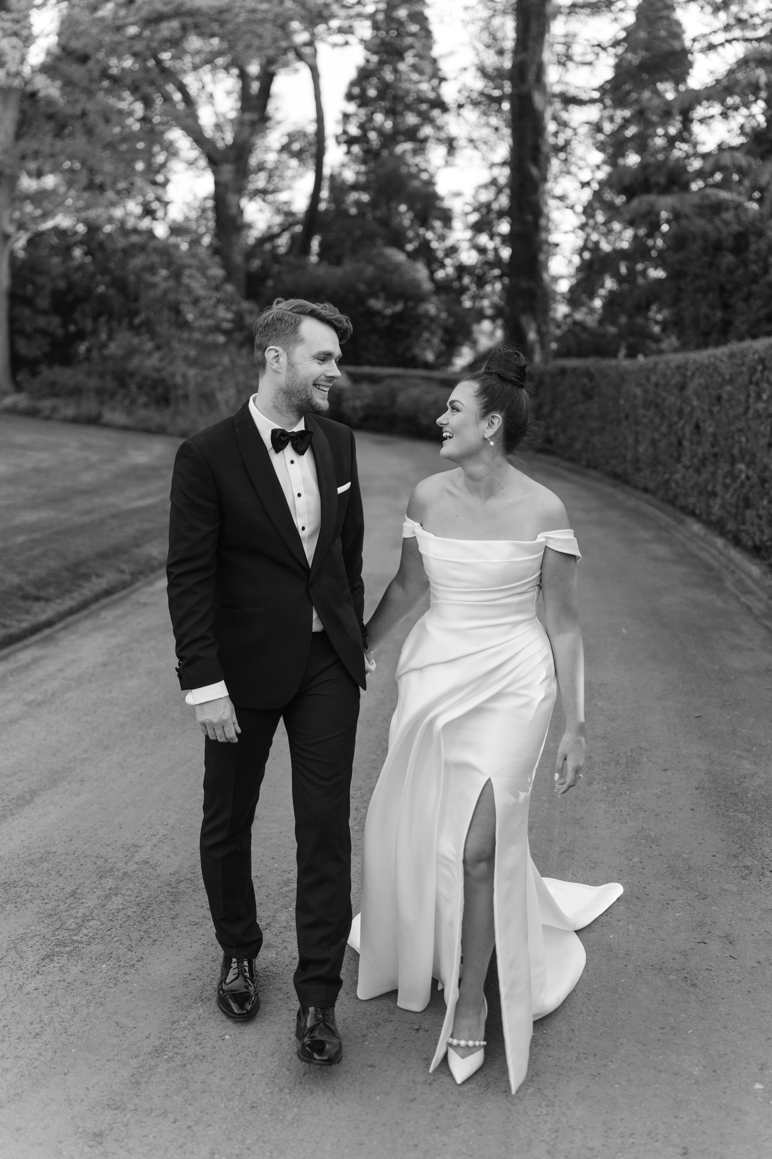 A black and white photo of a bride and groom walking on a path, surrounded by trees and hedges. The groom is in a tuxedo, and the bride is in a long, off-shoulder gown with a slit.