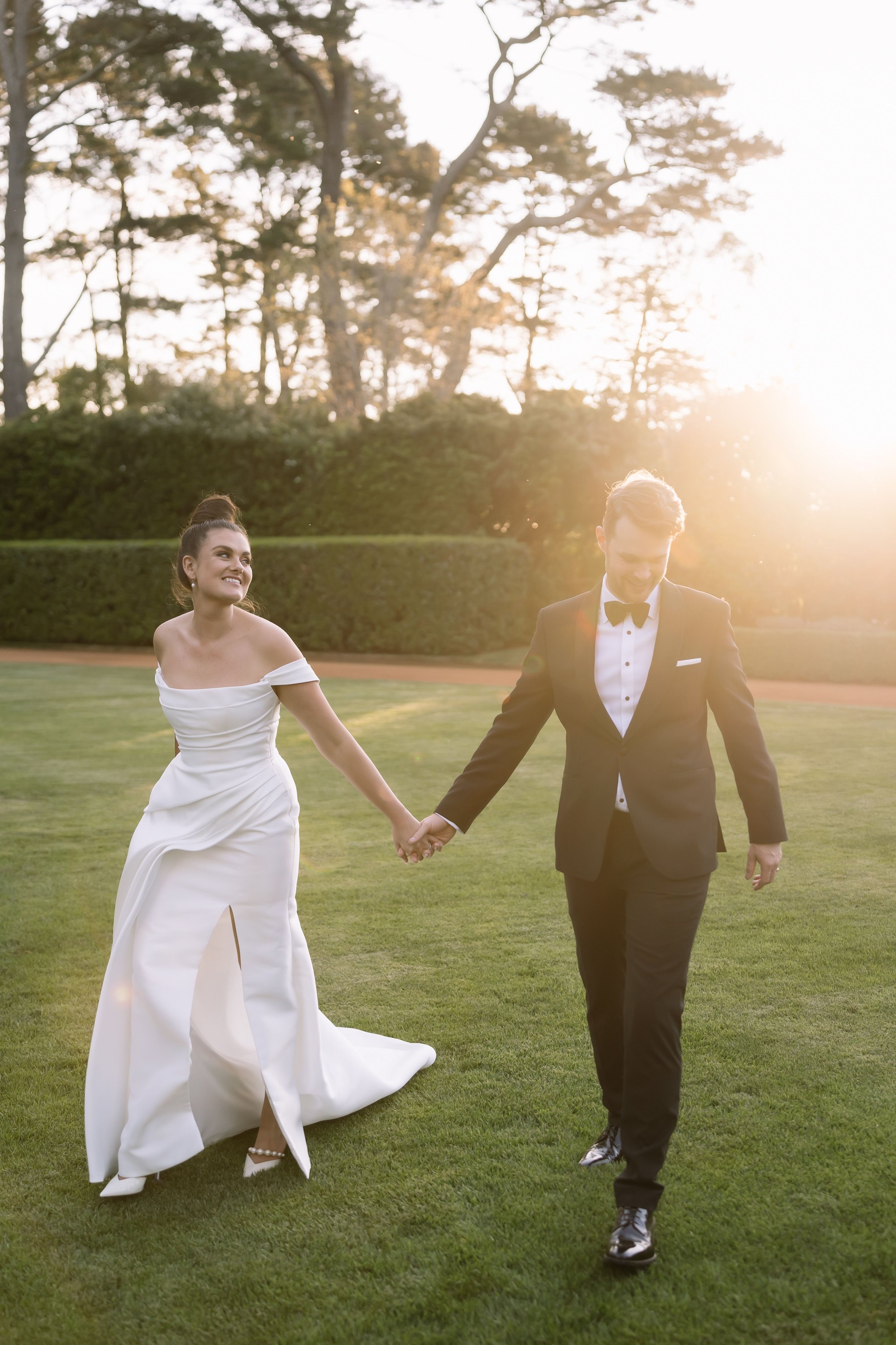 Bride in white gown and groom in black tuxedo walking hand in hand on a green lawn, backlit by the sun.