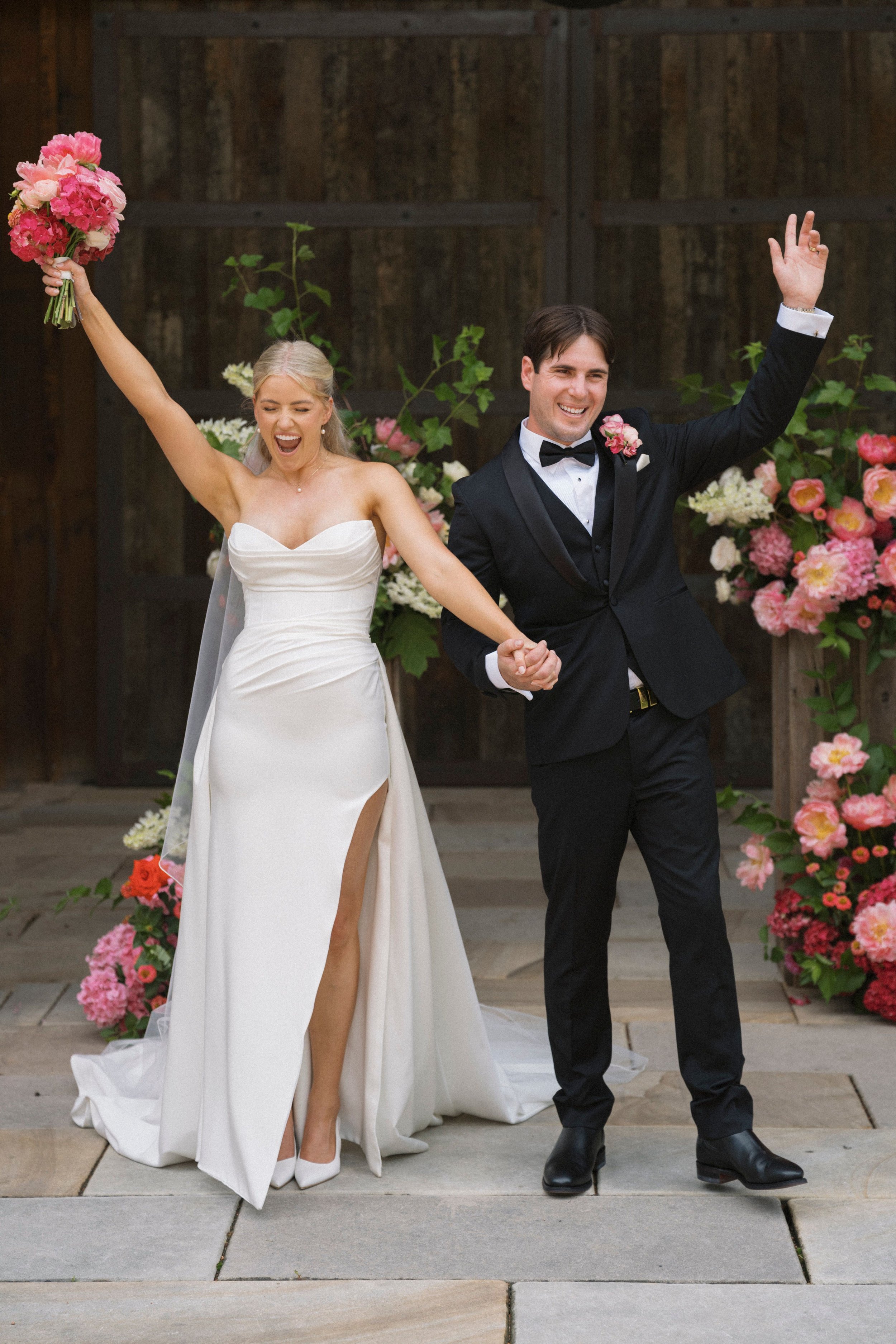 Bride and groom celebrating at a wedding, holding hands and raising arms joyfully, surrounded by colorful flowers.
