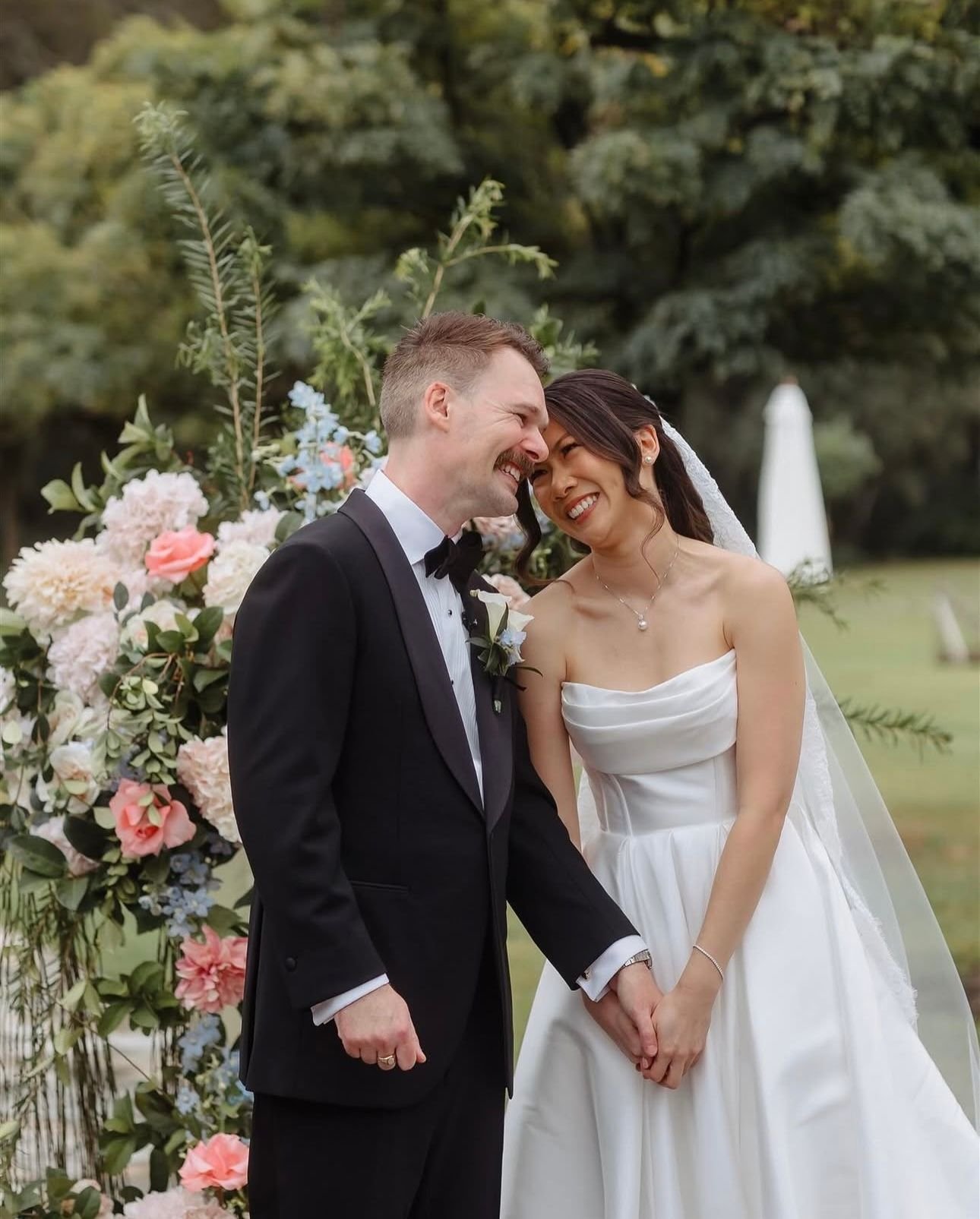 A bride and groom smiling and holding hands in an outdoor setting, surrounded by a floral arrangement, during their wedding.