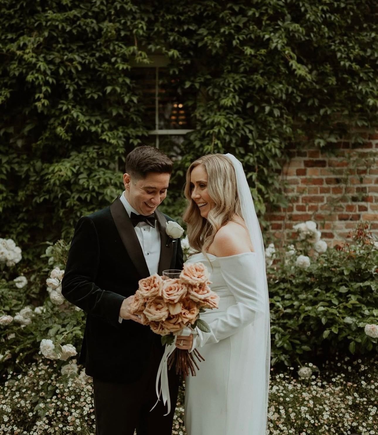 A bride and groom laughing in a garden, with the bride holding a bouquet of flowers. The background features ivy-covered brick walls and greenery.