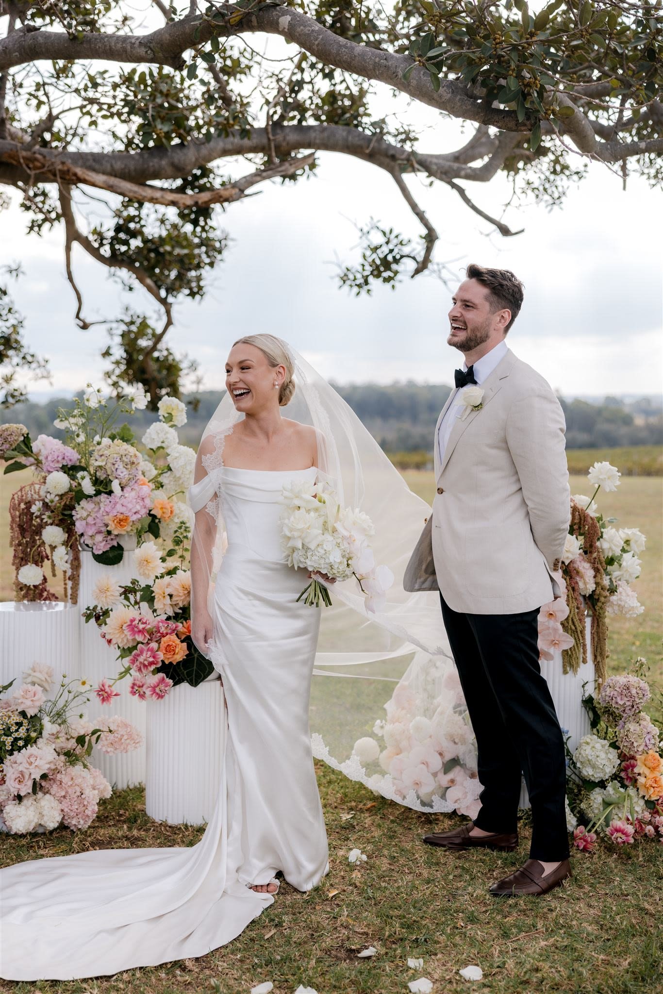 Bride and groom smiling, standing in front of floral decorations outdoors during wedding ceremony.