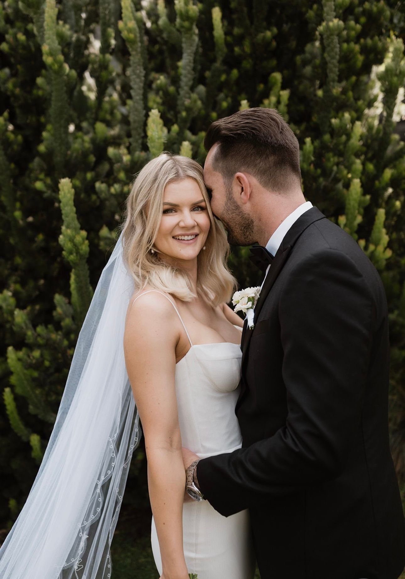 A bride and groom in formal attire with the groom wearing a black suit and the bride in a white dress with a veil, posing outdoors in front of green foliage, with the groom whispering to the bride who is smiling.
