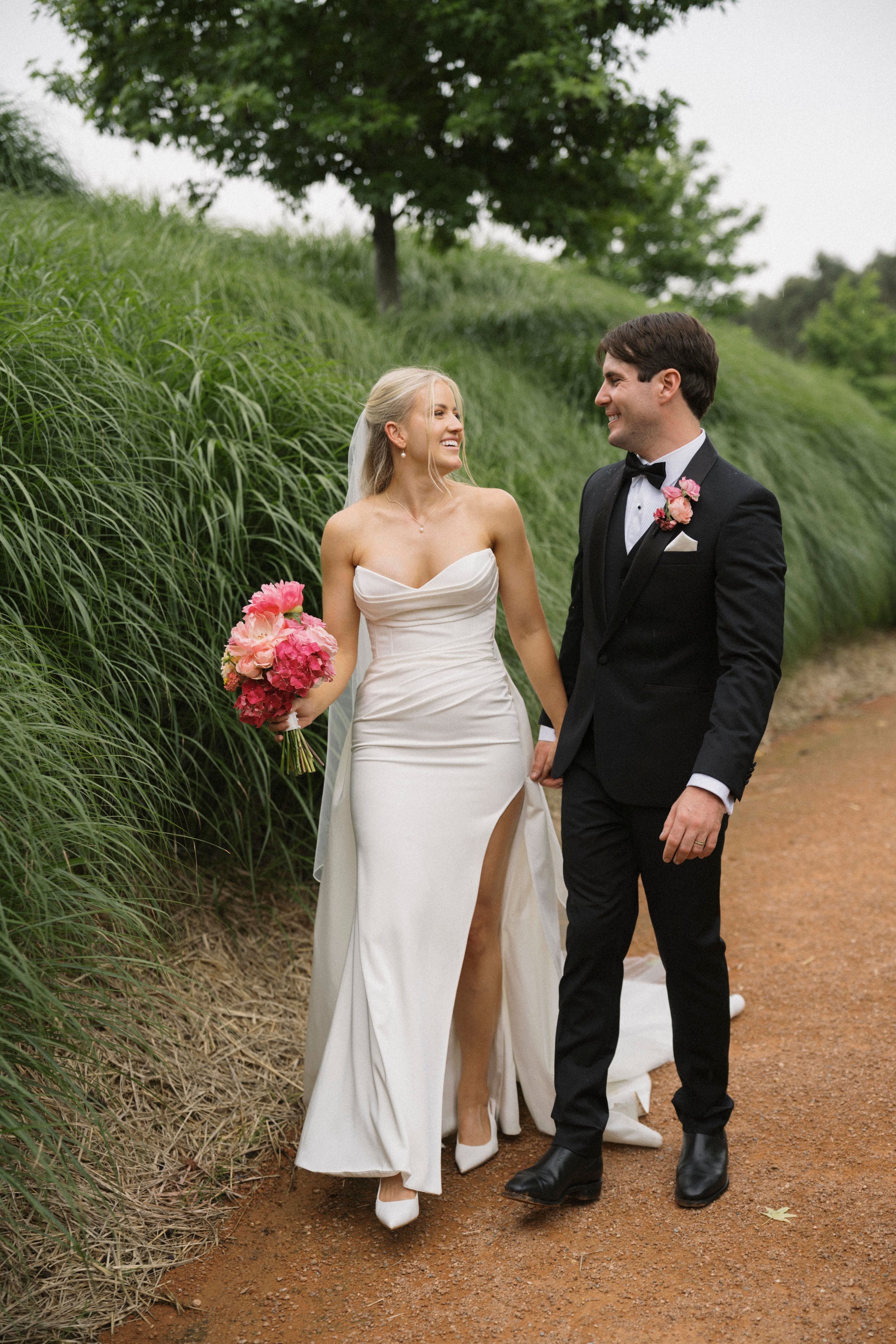 A bride in a white dress and a groom in a black tuxedo walking on a path surrounded by greenery, with the bride holding a bouquet of pink flowers.