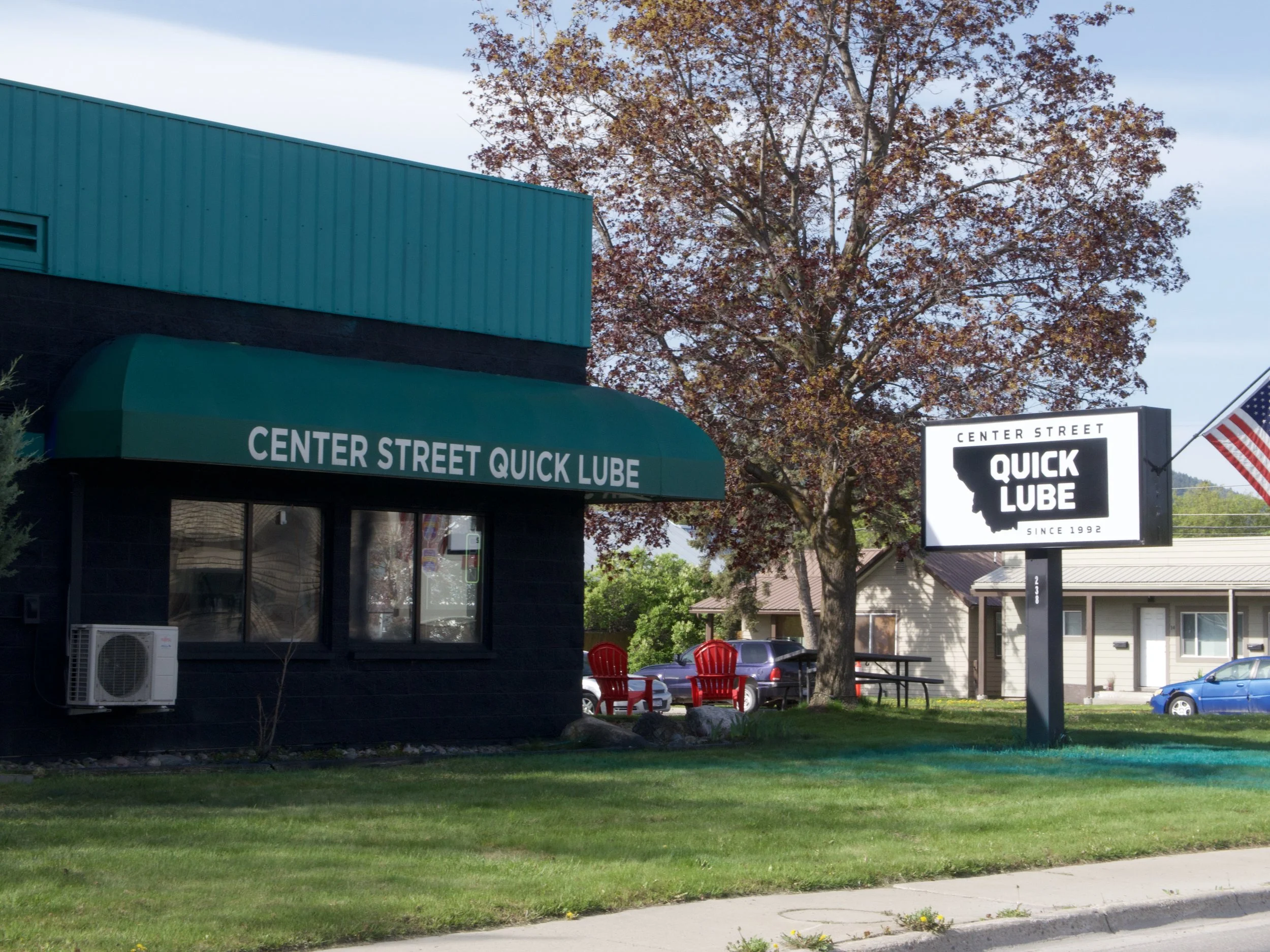 A street scene with a building featuring a green awning that says 'CENTER STREET QUICK LUBE,' a parking lot with cars, two red chairs, a large tree with autumn-colored leaves, and some signage including a black and white sign for 'Quick Lube' and an American flag.