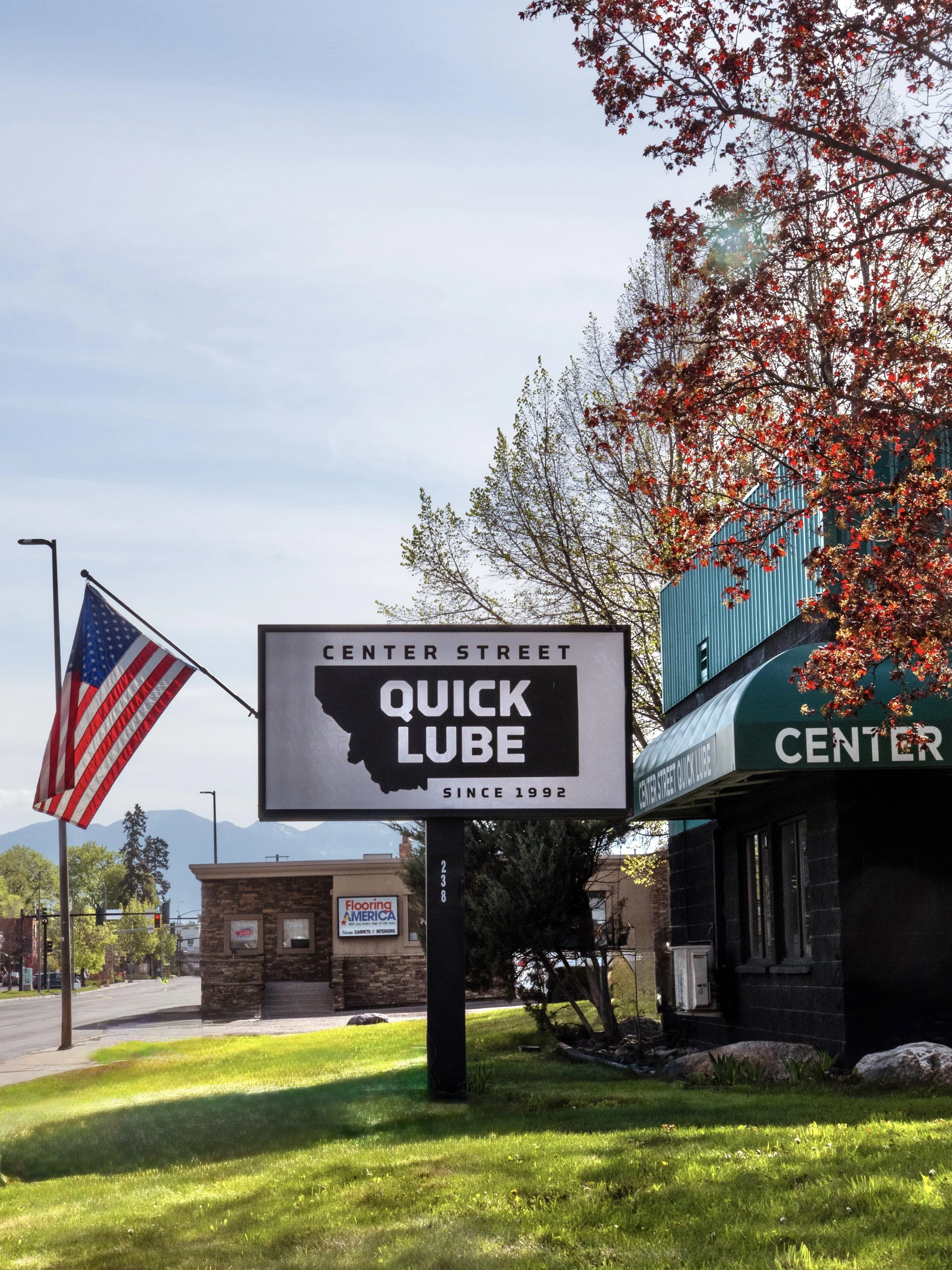 Street scene with a sign for 'Center Street Quick Lube' and American flags on poles, trees with red leaves, a blue building, and distant mountains in the background.