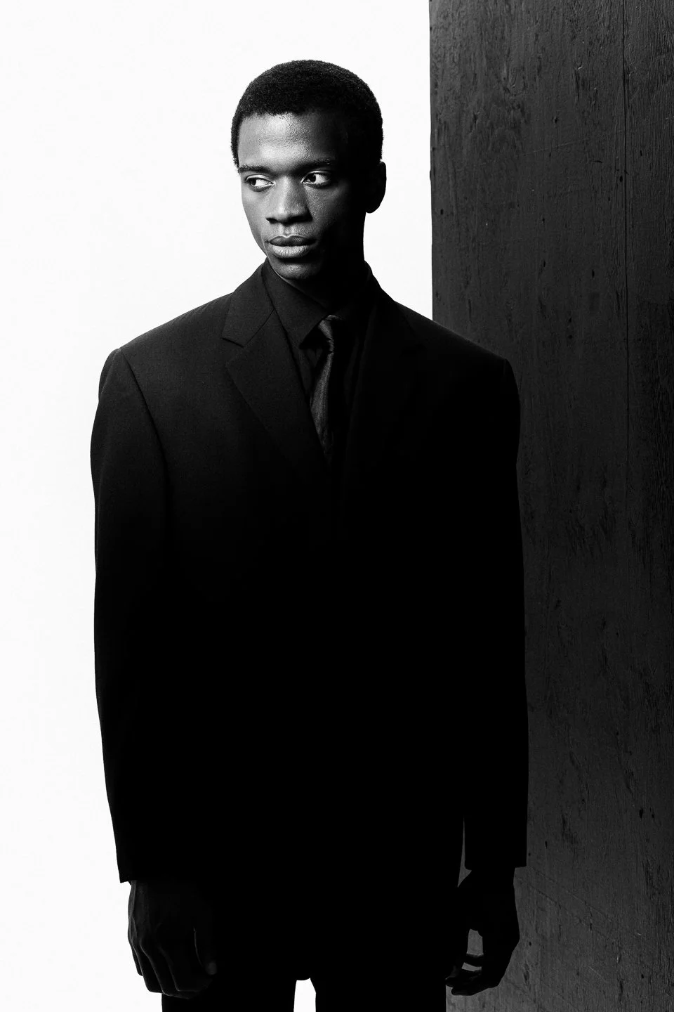 Un homme noir en costume noir, regardant vers la gauche, en noir et blanc, debout contre un mur foncé à droite et un fond blanc à gauche.