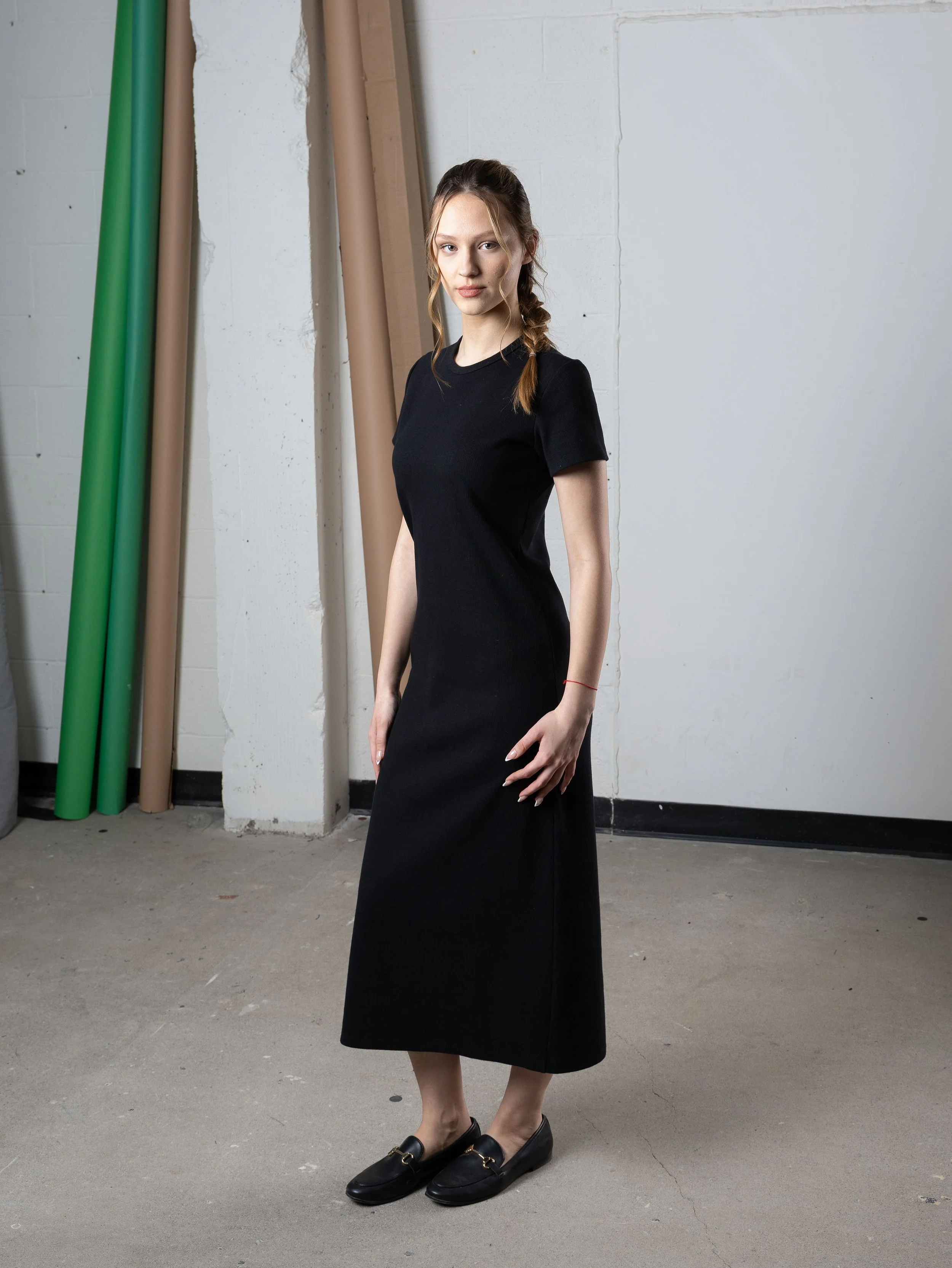 Une jeune femme portant une robe noire longue et des mocassins noirs, se tenant dans un espace industriel avec des tuyaux verts et beiges en toile de fond.