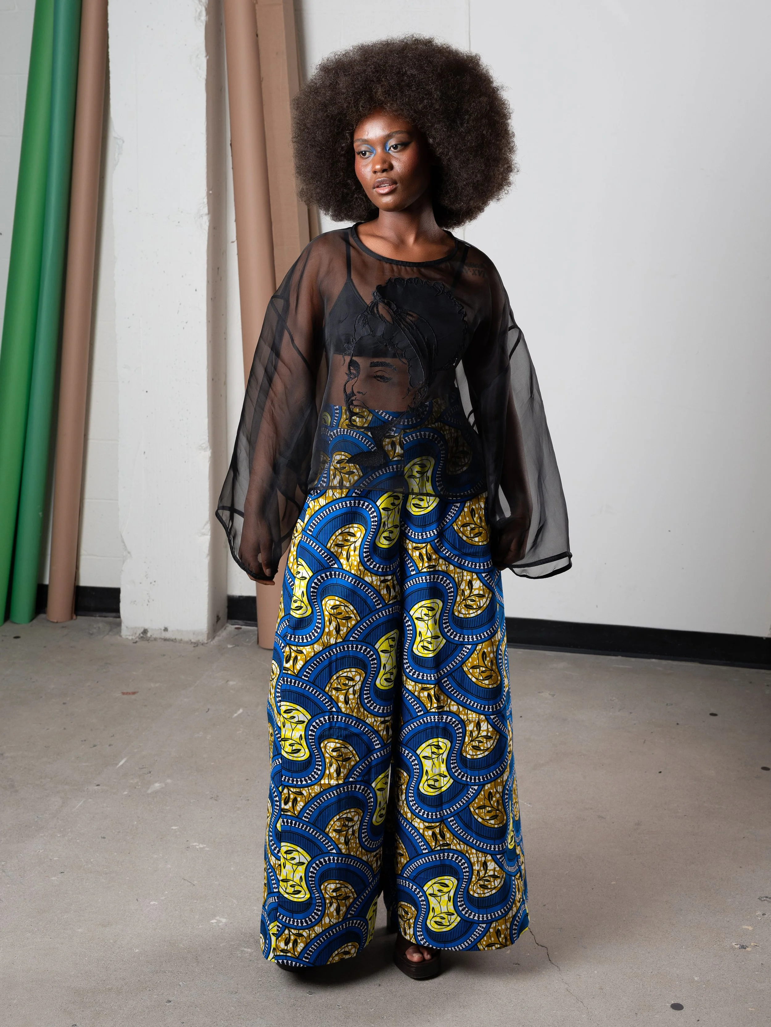 Femme portant un haut en tulle noir avec une illustration d'une femme en print sur le devant et un pantalon ample à motif africain coloré en bleu, jaune et noir.