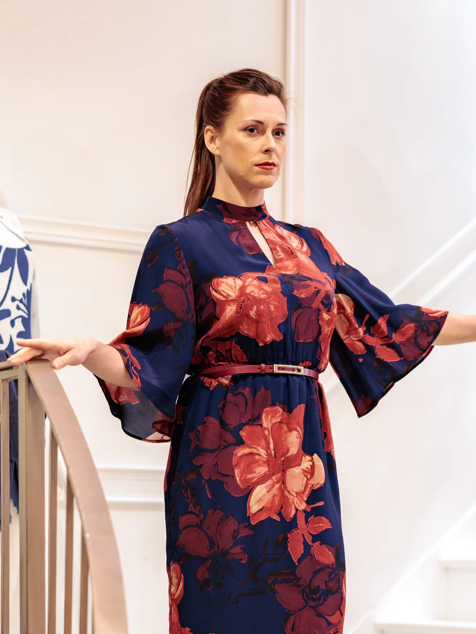 Une femme avec des cheveux longs bruns, portant une robe à fleurs rouge et bleu avec une ceinture, se tient près d'une rampe d'escalier blanche à l'intérieur d'une maison.