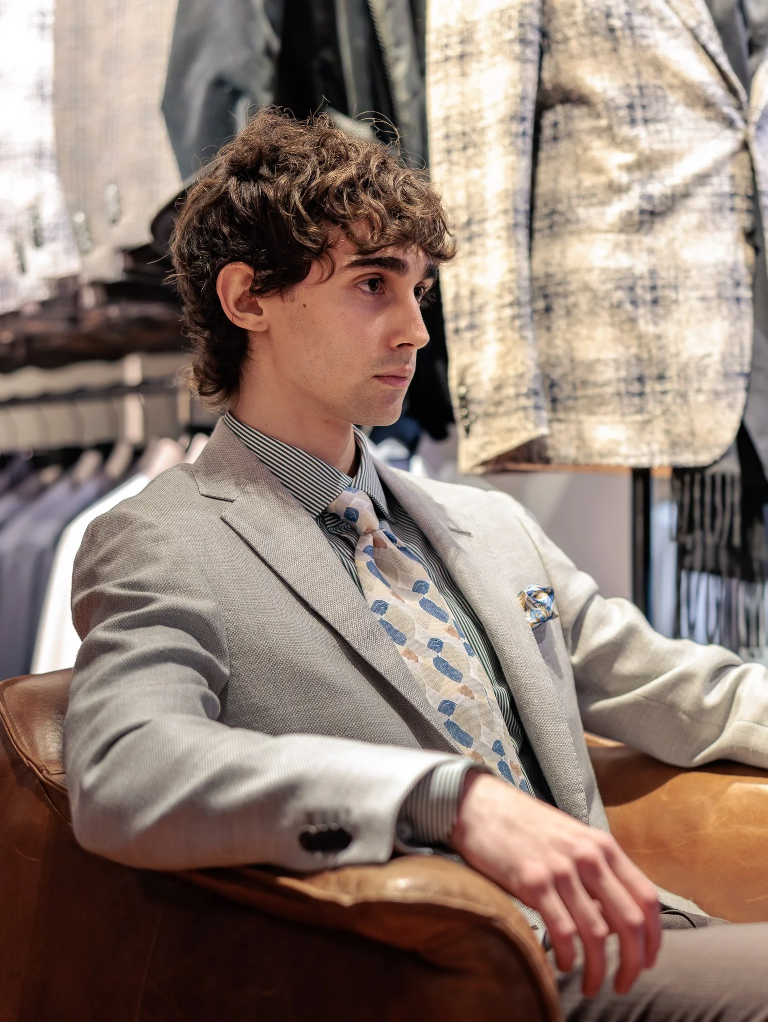 Un homme jeune assis dans un magasin ou un bureau, portant un costume crème, une chemise à rayures et une cravate avec un motif de forme arquée, assis sur un fauteuil en cuir marron, avec des vêtements suspendus en arrière-plan.