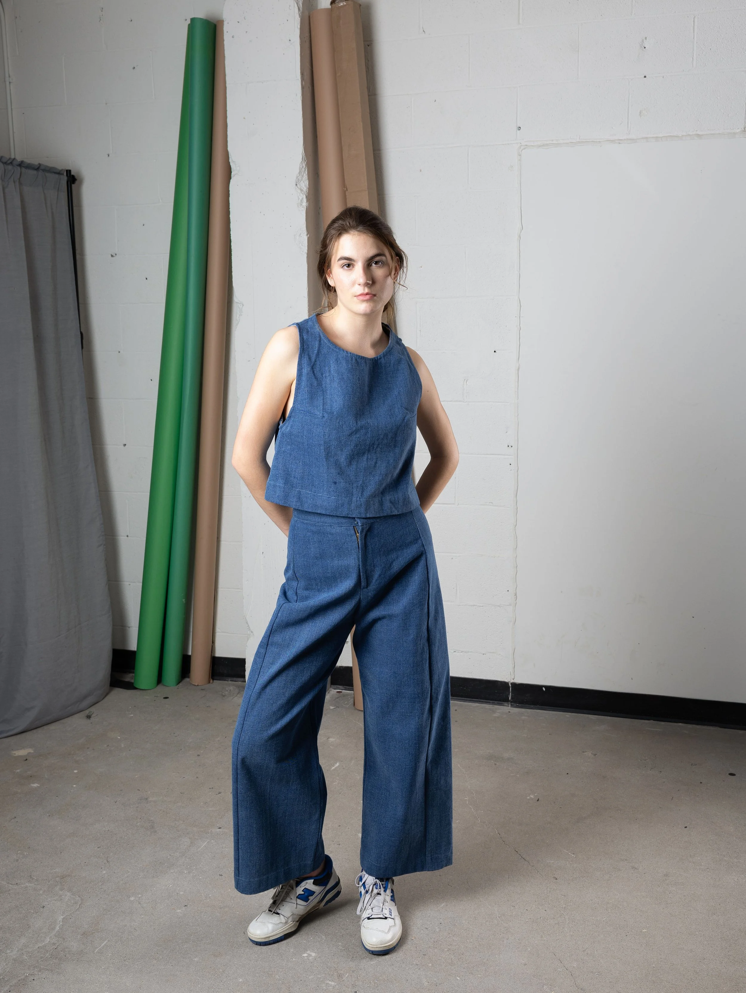 Jeune femme debout dans un espace industriel, portant une tenue en denim et des baskets, avec des rouleaux de papier derrière elle.