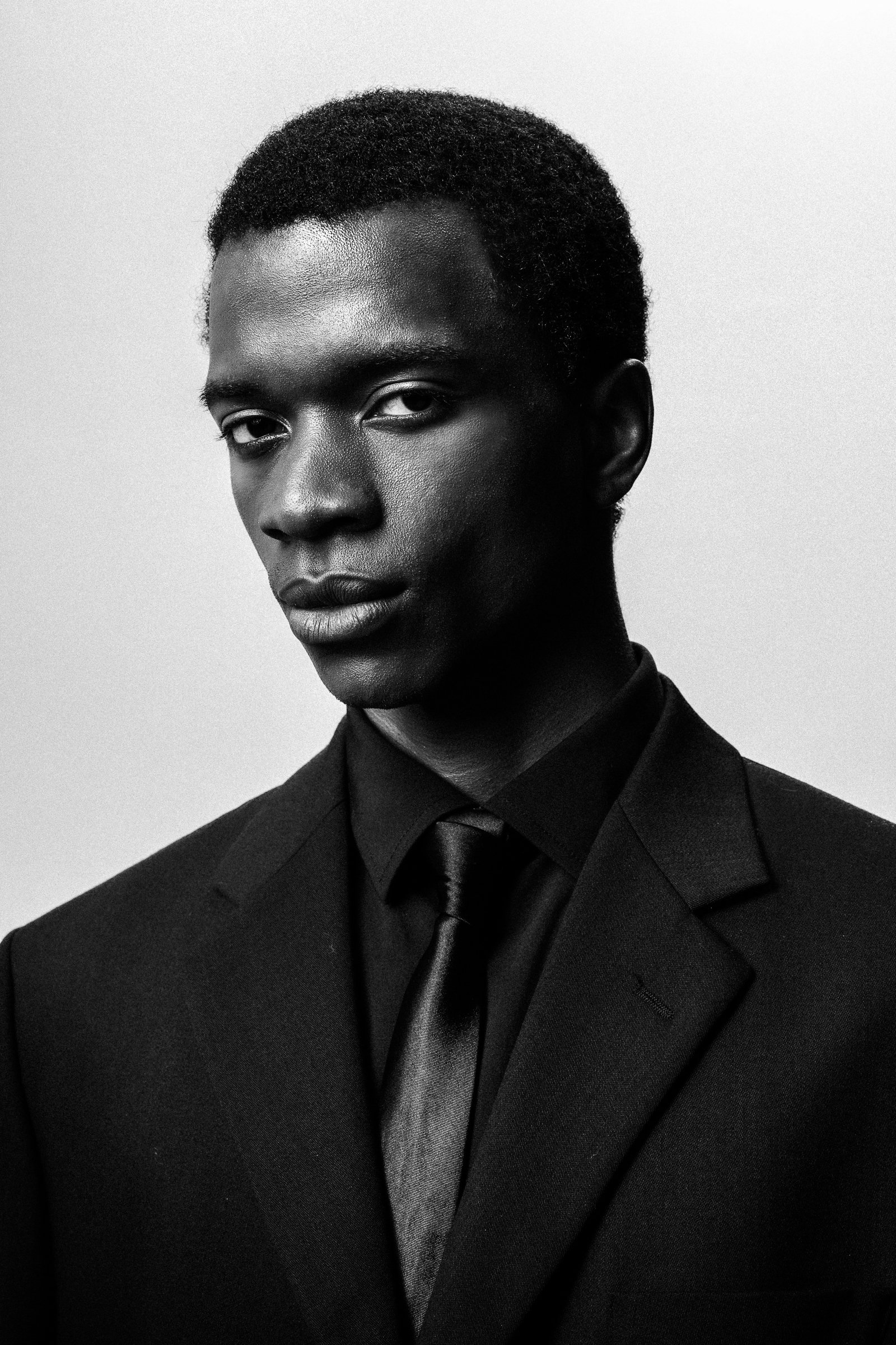 Portrait en noir et blanc d'un jeune homme portant un costume noir, chemise et cravate, regardant fixé vers l'objectif.