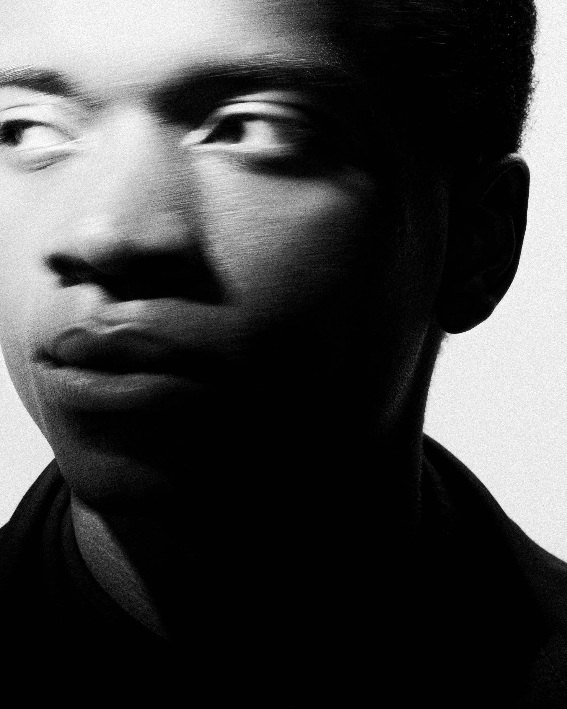 Portrait en noir et blanc d'une personne avec un regard vers la gauche, sourcils épais et lèvres fermées, avec éclat de lumière sur le visage.