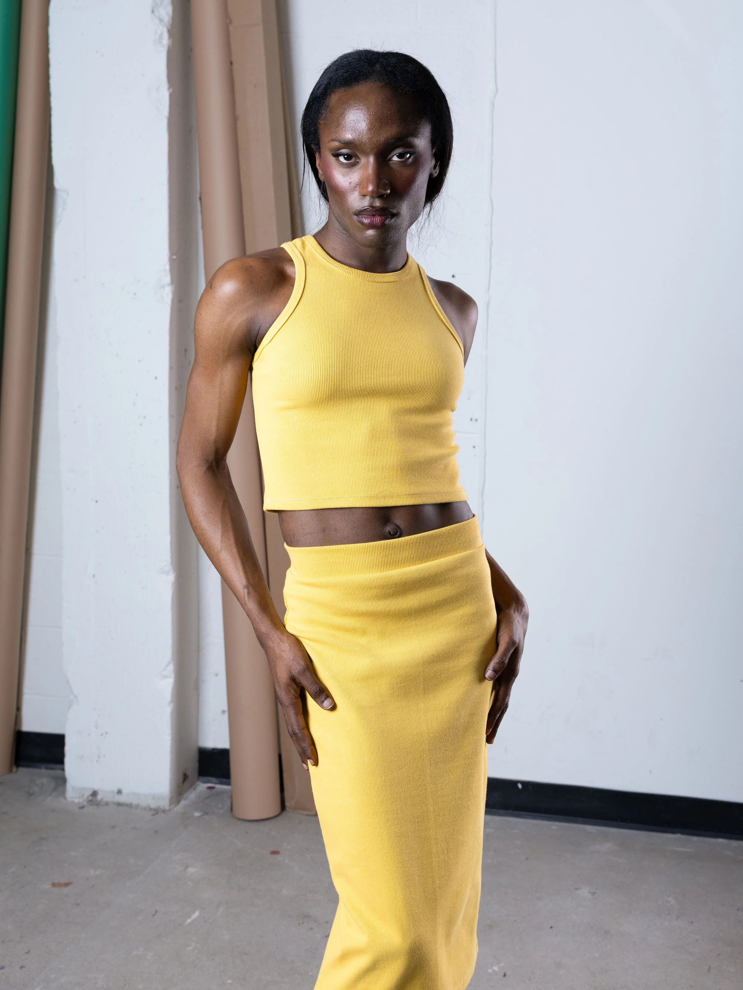 Une femme portant un haut et une jupe jaunes, posant dans un studio avec un mur blanc en arrière-plan.