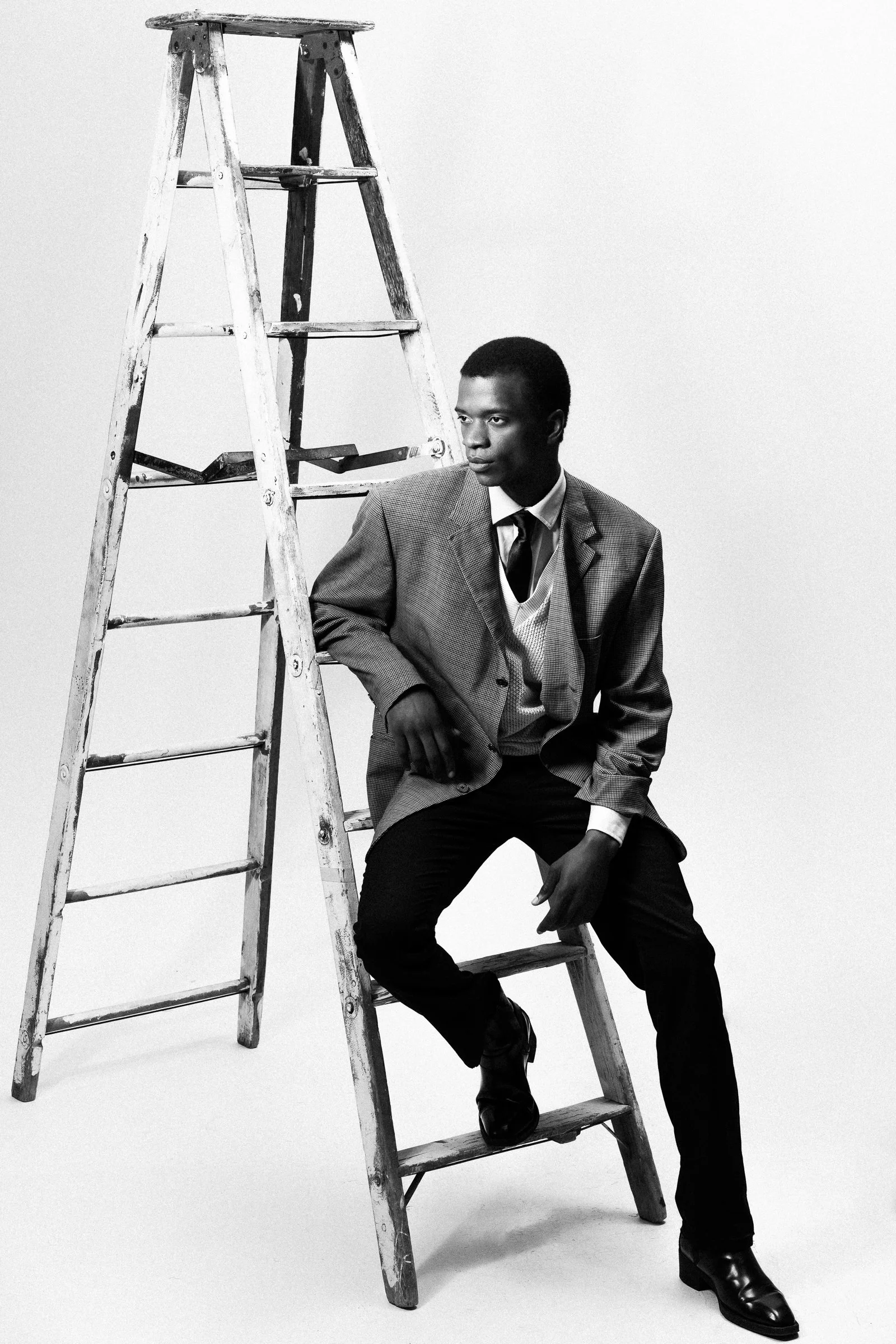 Un homme portant un costume, assis sur une échelle en bois, en noir et blanc.