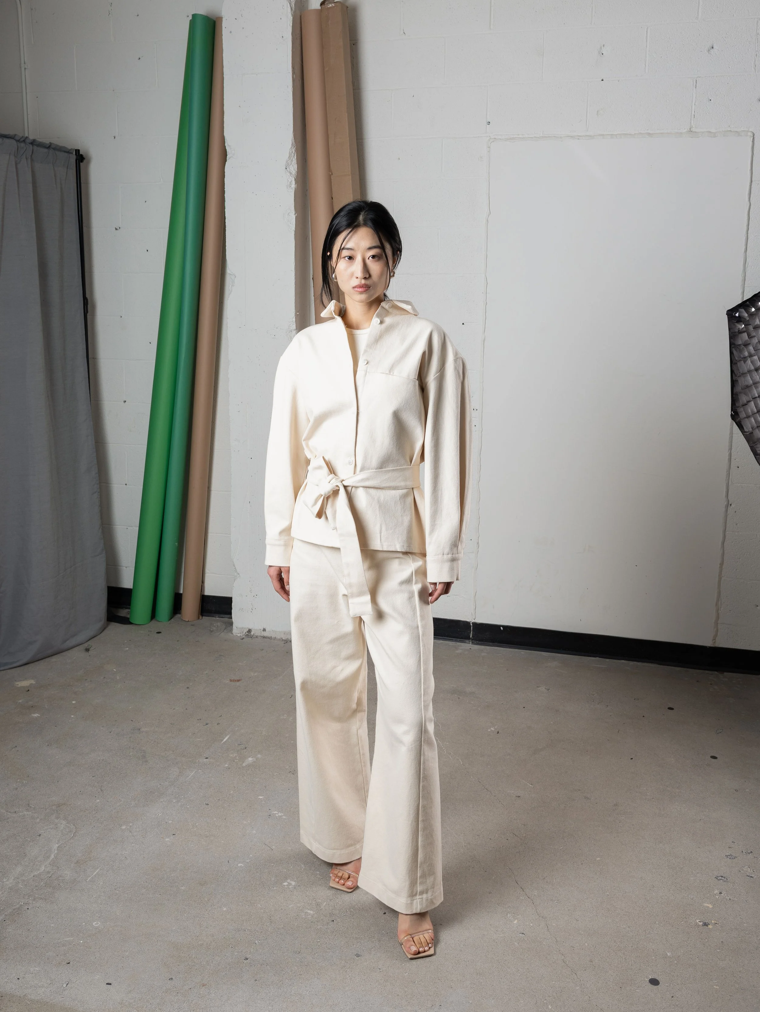 Une femme portant une tenue beige, debout dans un studio avec des rouleaux de papier en arrière-plan et un éclairage de studio à gauche.