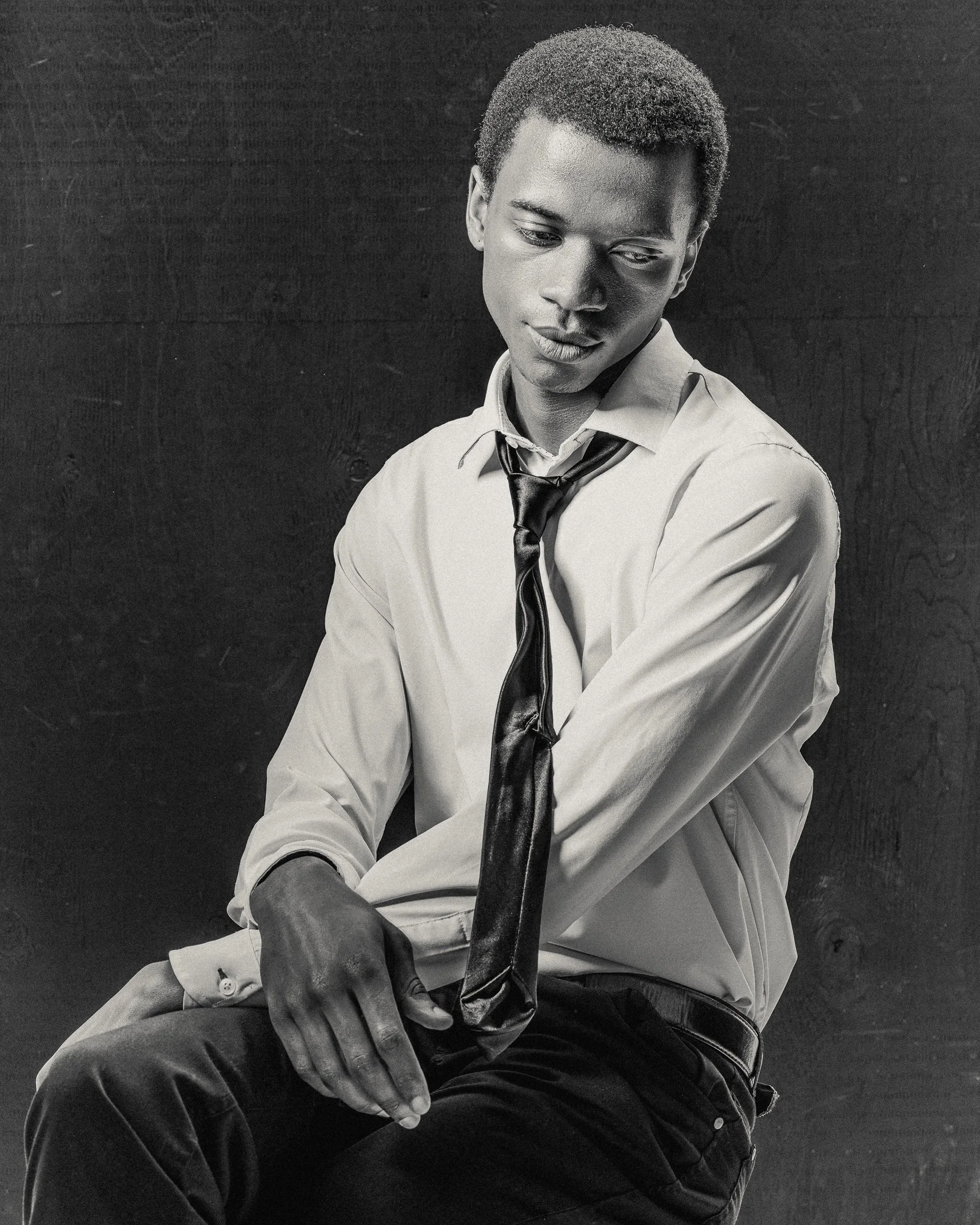 Jeune homme en chemise blanche et cravate noire, assis contre un fond sombre, regardant vers le bas.