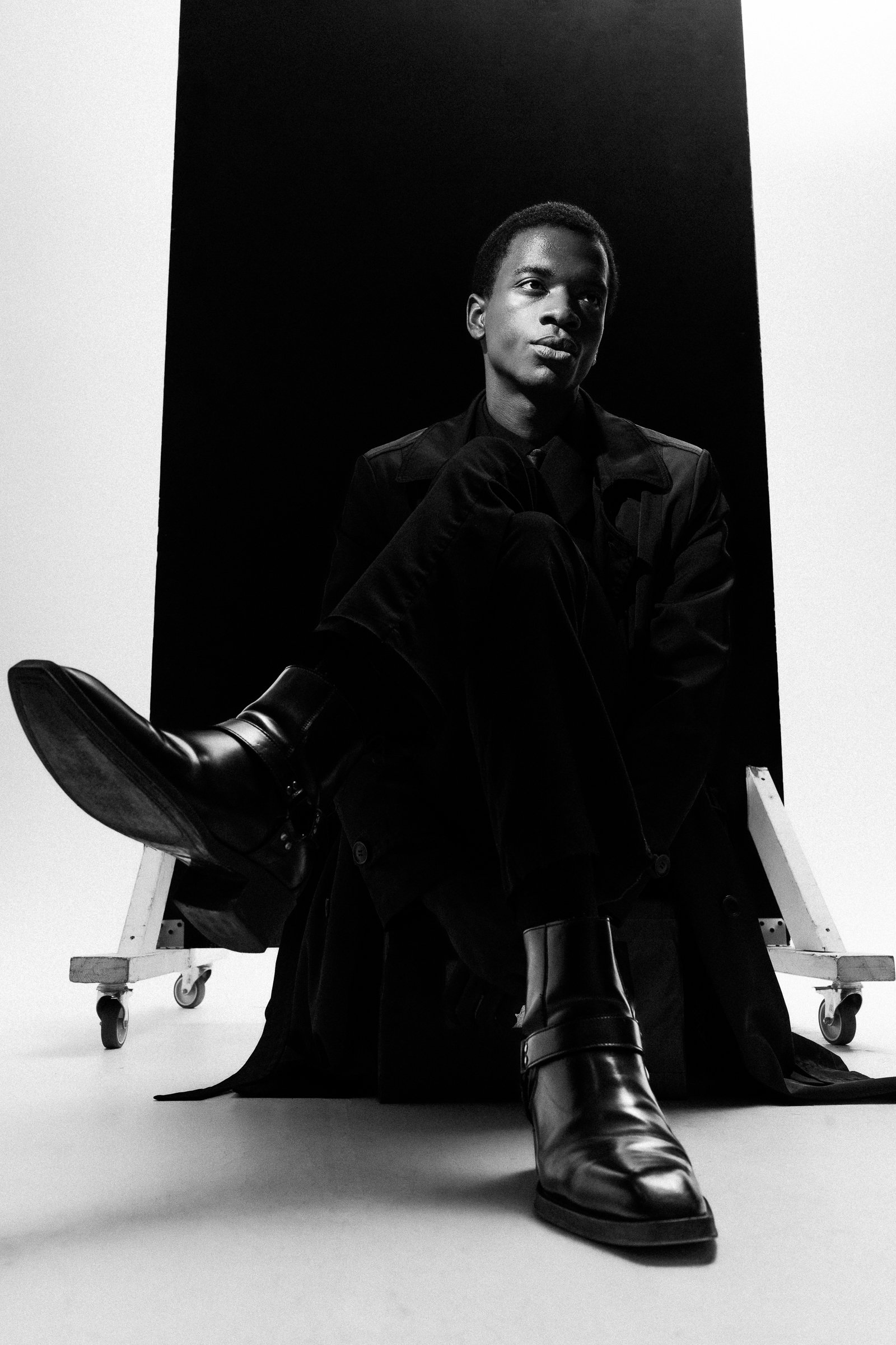 Jeune homme assis devant un fond noir, en tenue sombre, portant des bottes en cuir, dans une séance photo en noir et blanc.