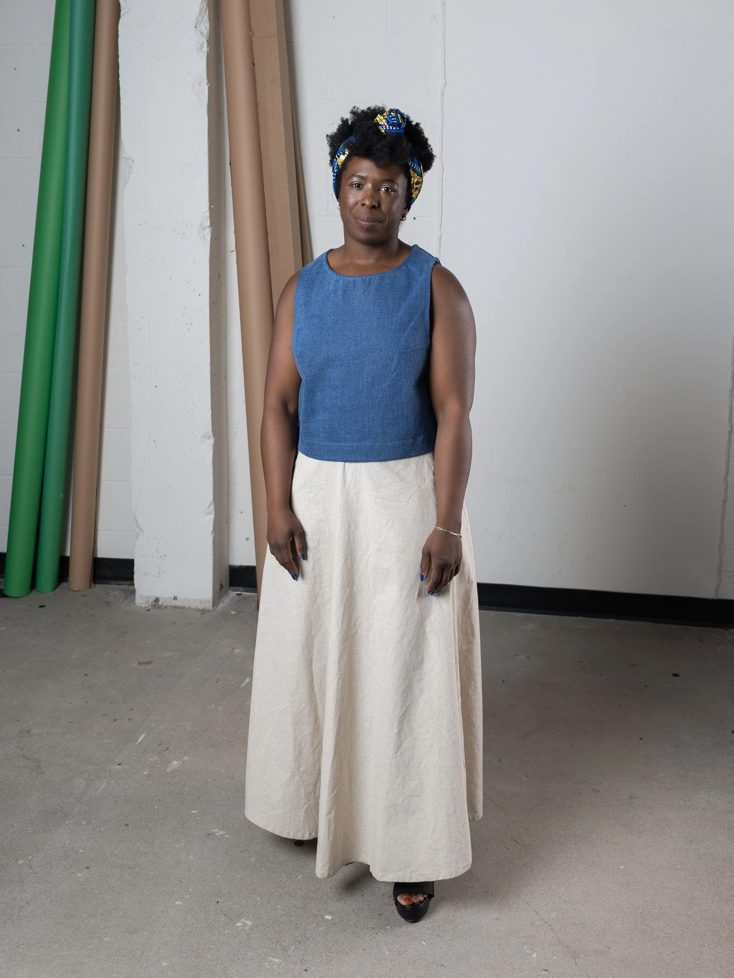 Une femme debout dans une pièce, portant un haut sans manches bleu et une longue jupe beige, avec un bandeau coloré dans les cheveux.