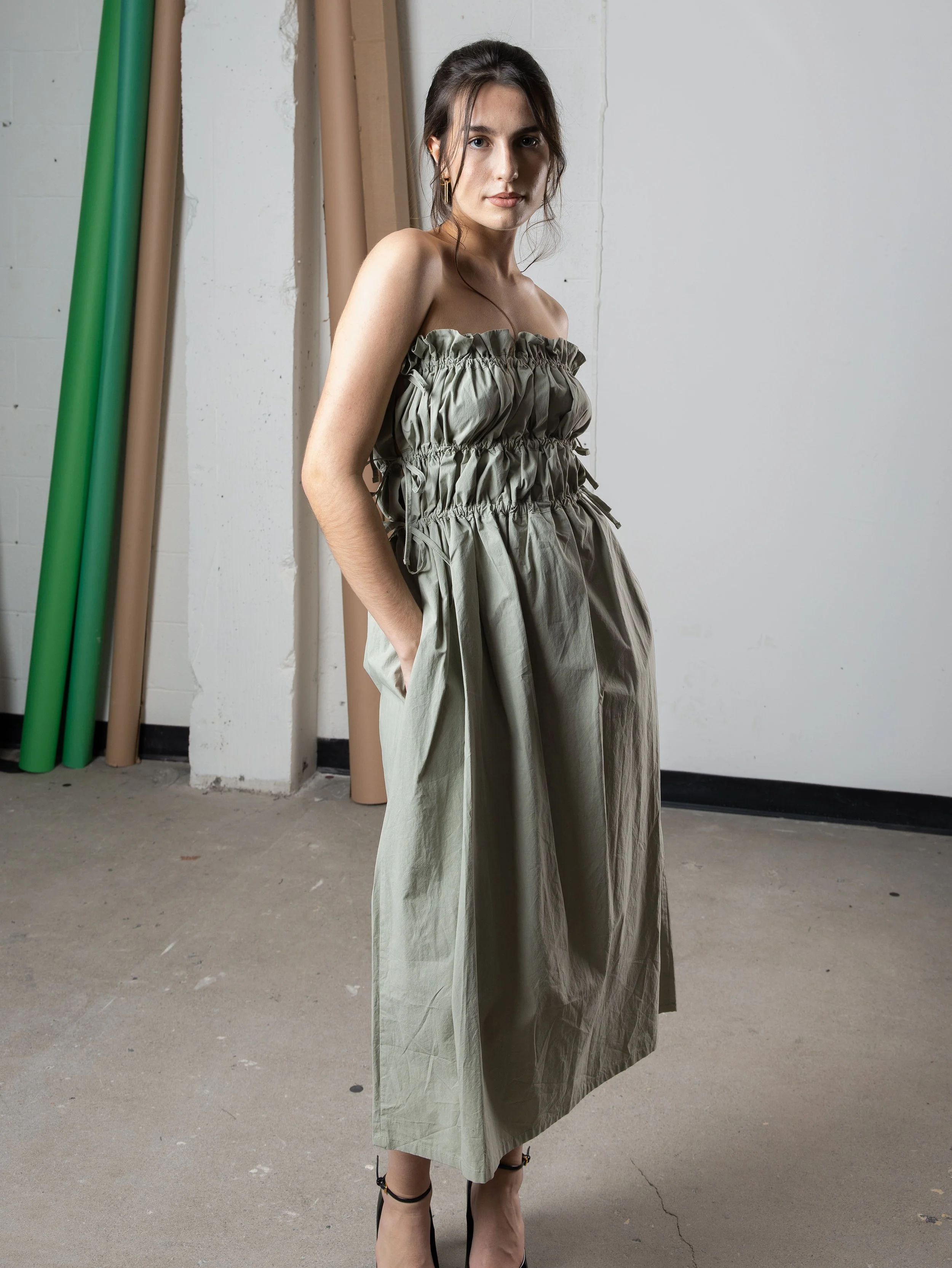 Jeune femme debout dans un atelier en portant une robe ample de couleur vert olive et des talons noirs.