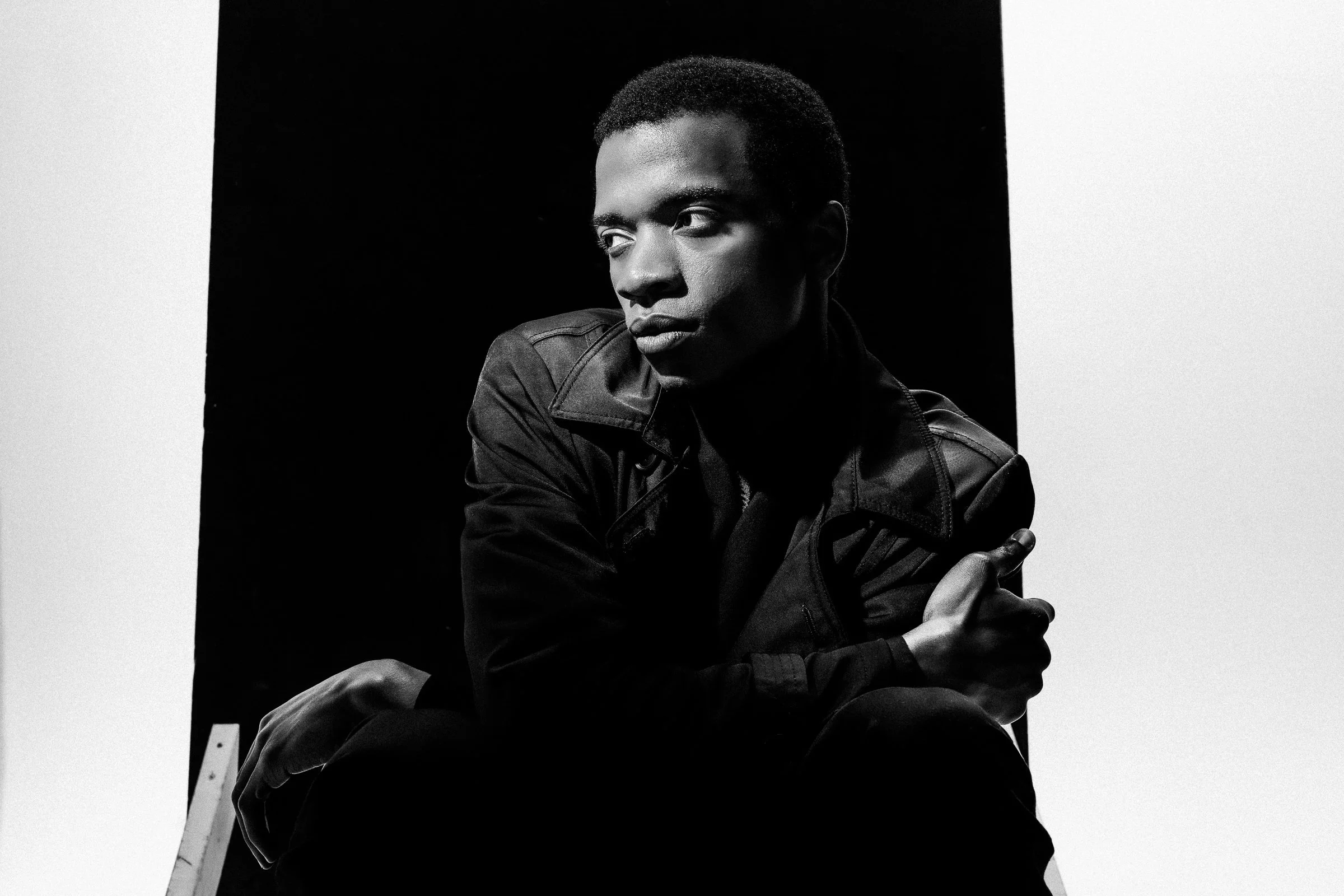 Un portrait en noir et blanc d'un homme jeune portant une veste en cuir, assis contre un fond noir et blanc, regardant vers la gauche.