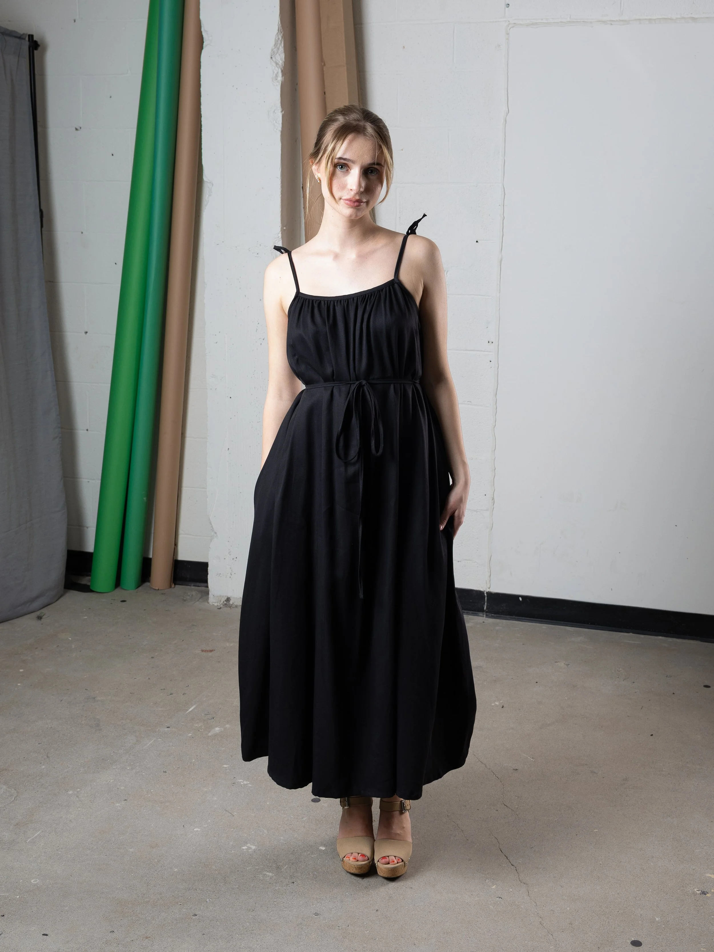 Jeune femme portant une robe noire longue à baillettes, avec des talons beiges, portant des cheveux mi-longs et bouclés, se tenant dans un espace intérieur avec des rouleaux de fond en plastique vert et beige en arrière-plan.