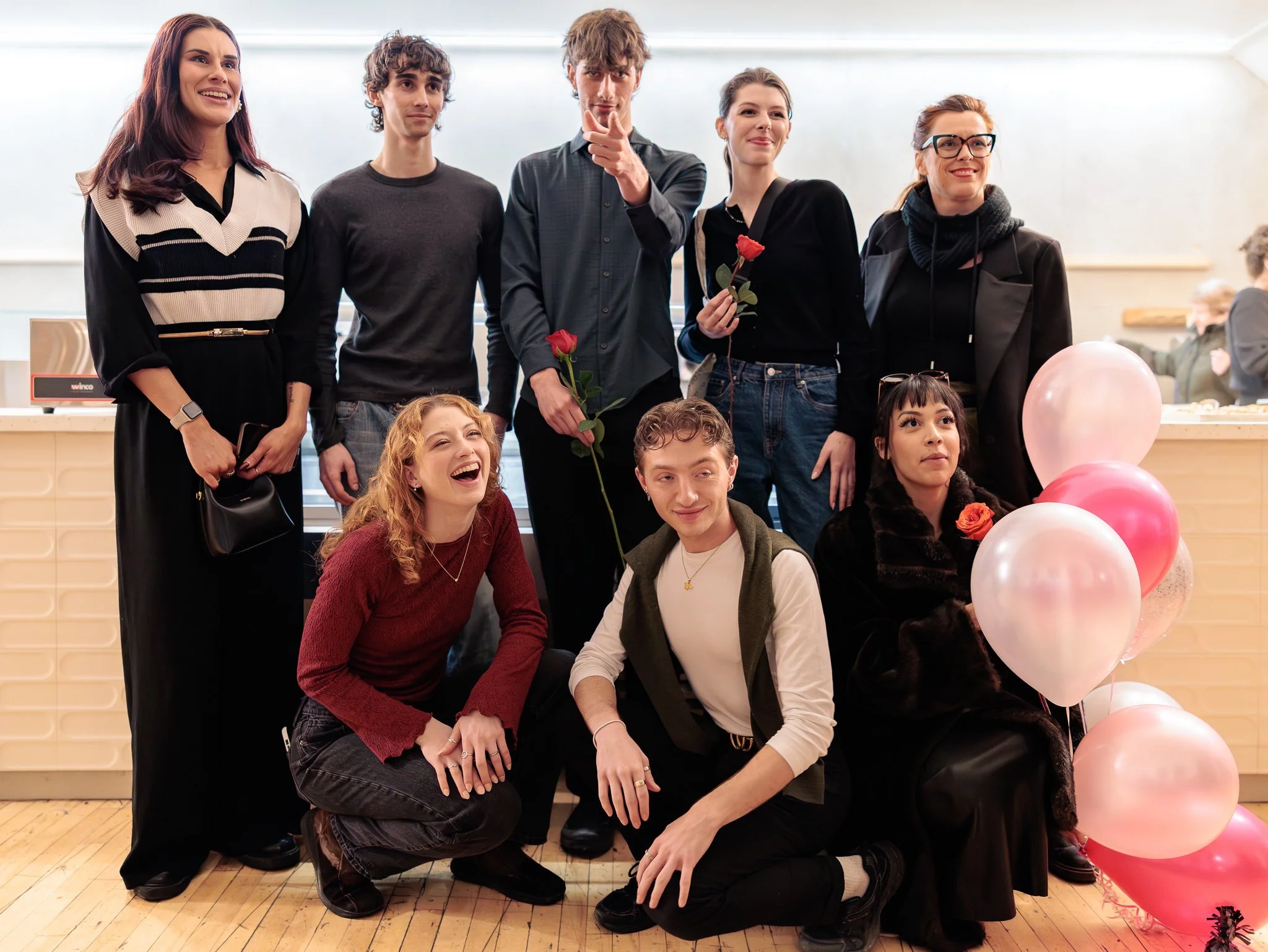 Groupe de neuf jeunes adultes posant pour une photo à l'intérieur, certains tenant des roses et entourés de ballons roses et blancs, lors d'une fête ou célébration.