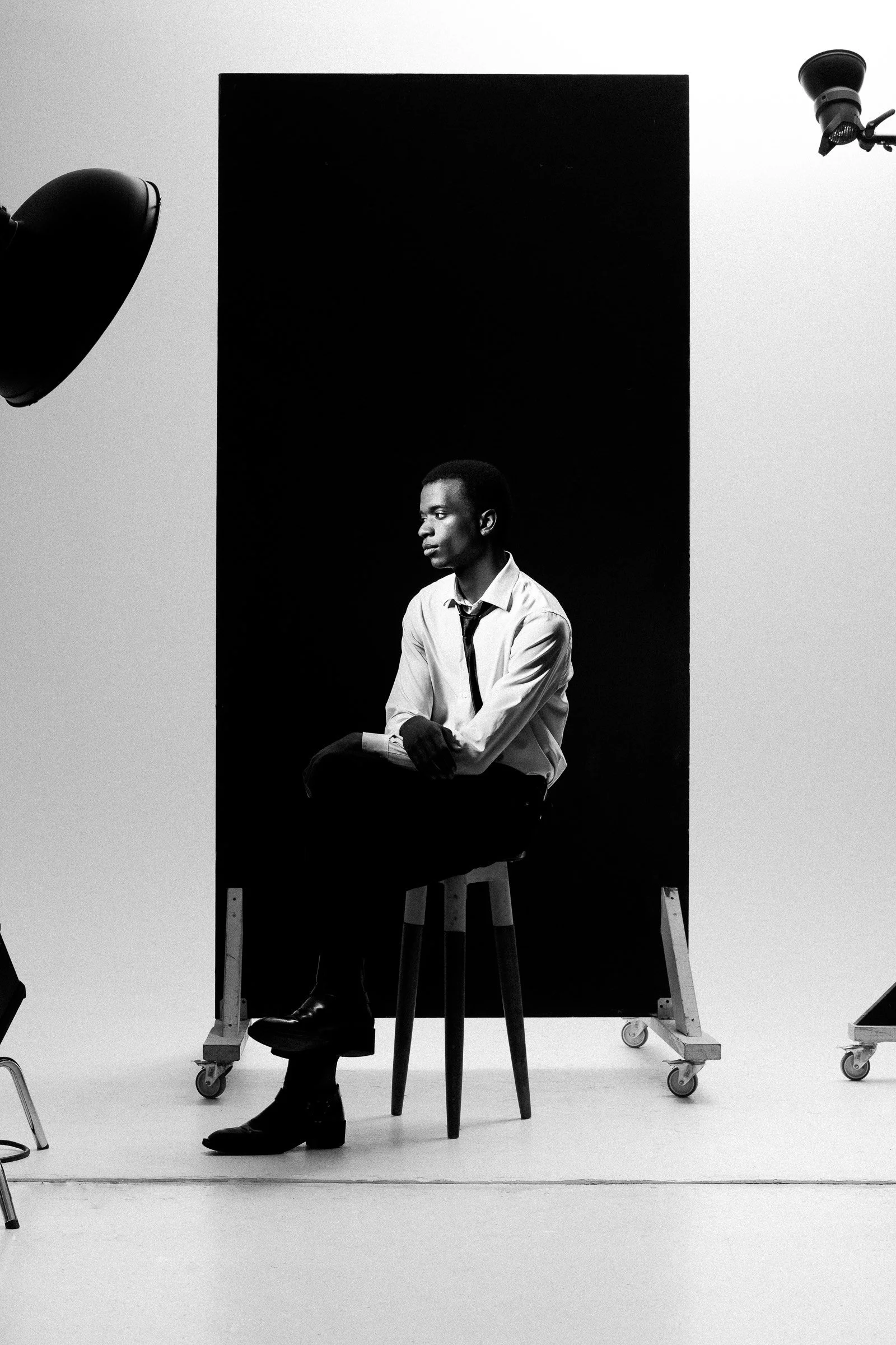 Un homme assis sur une chaise en studio photo, en noir et blanc, avec un fond sombre et des équipements d'éclairage