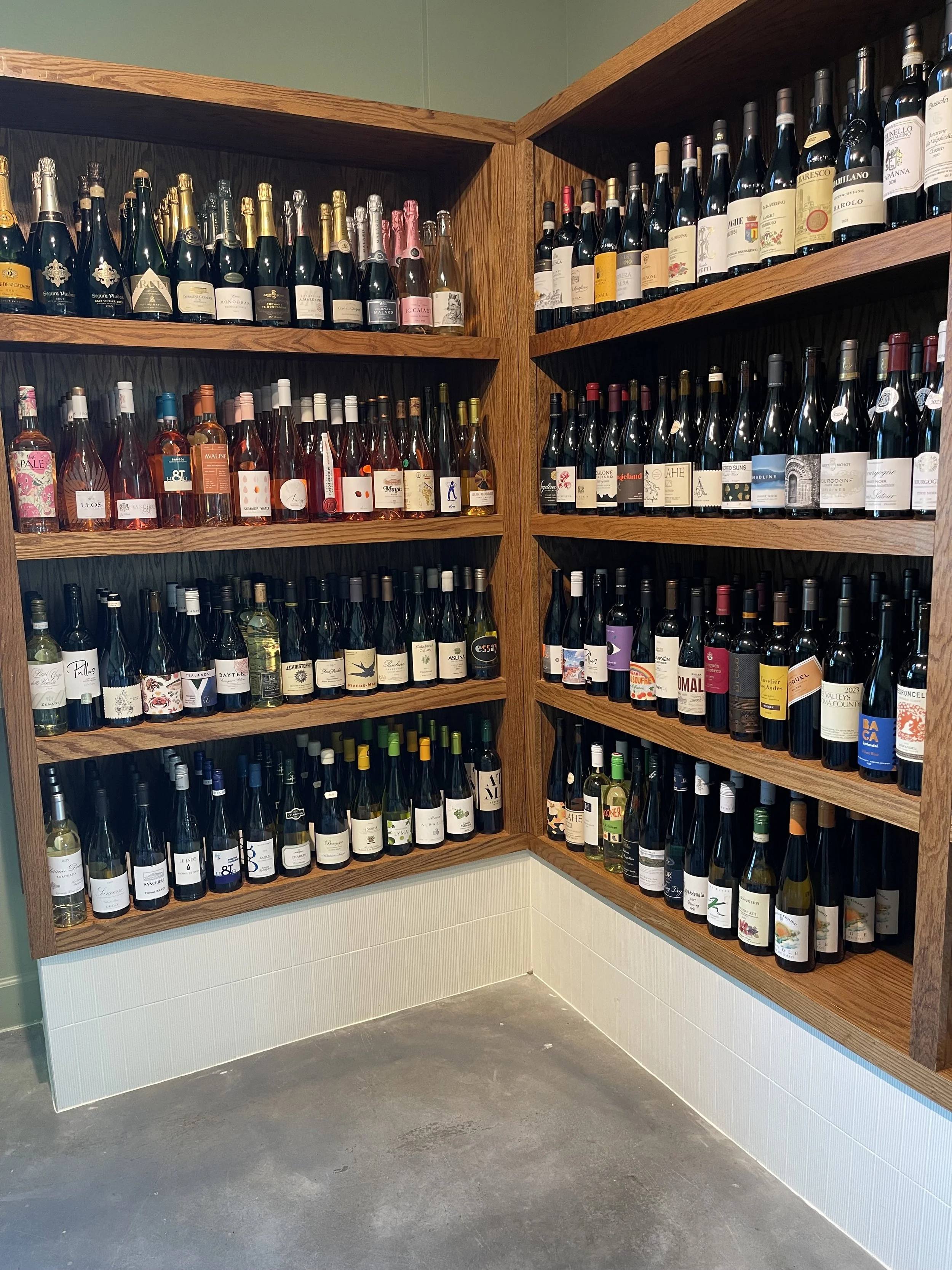 Wall of wine bottles displayed on wooden shelves in a wine cellar or store, with various labels and colors.