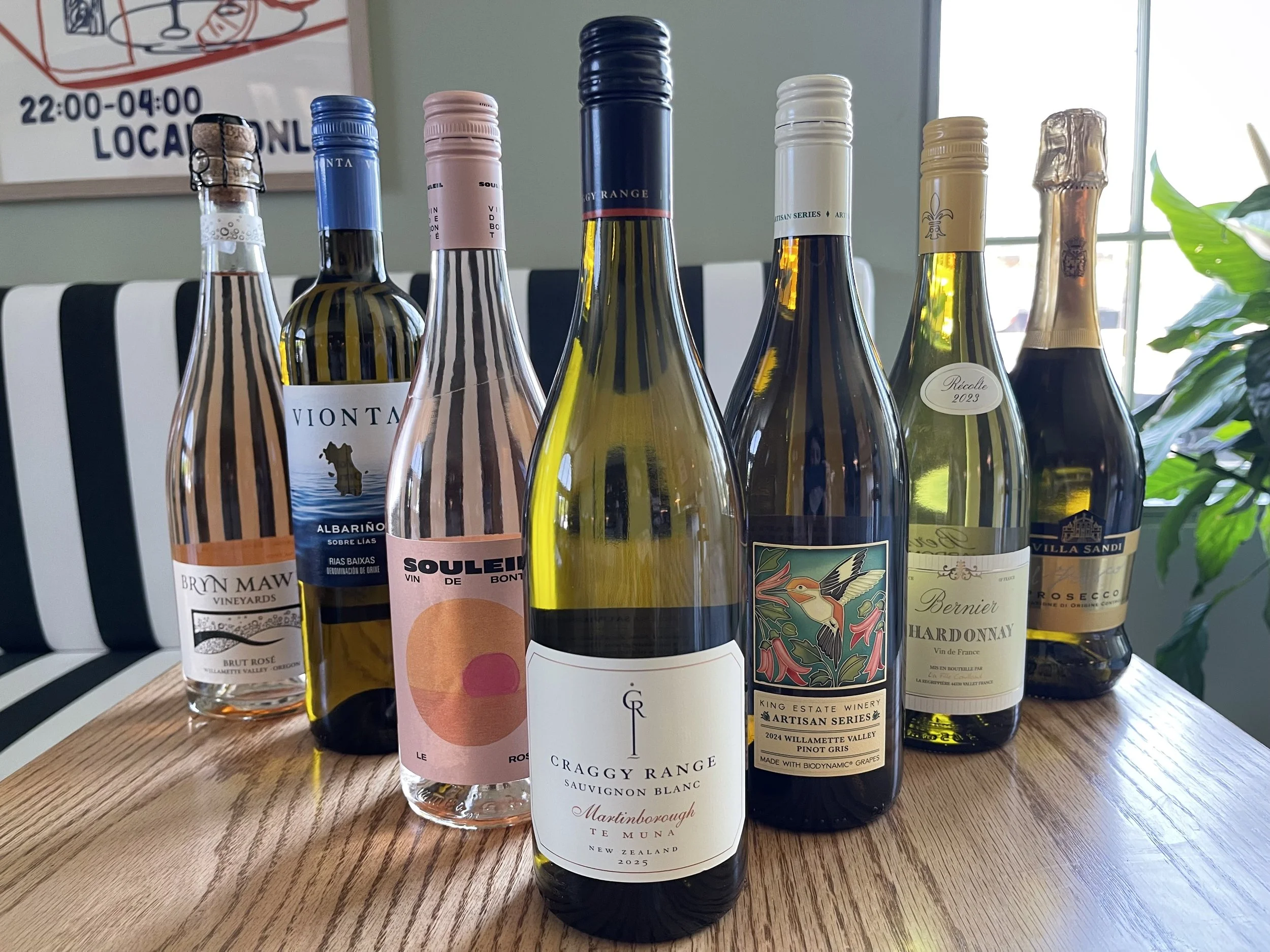 A row of various wine bottles with colorful labels on a wooden table, some with metallic foil capsules, and a background with a striped black and white chair, a tree, and a window.
