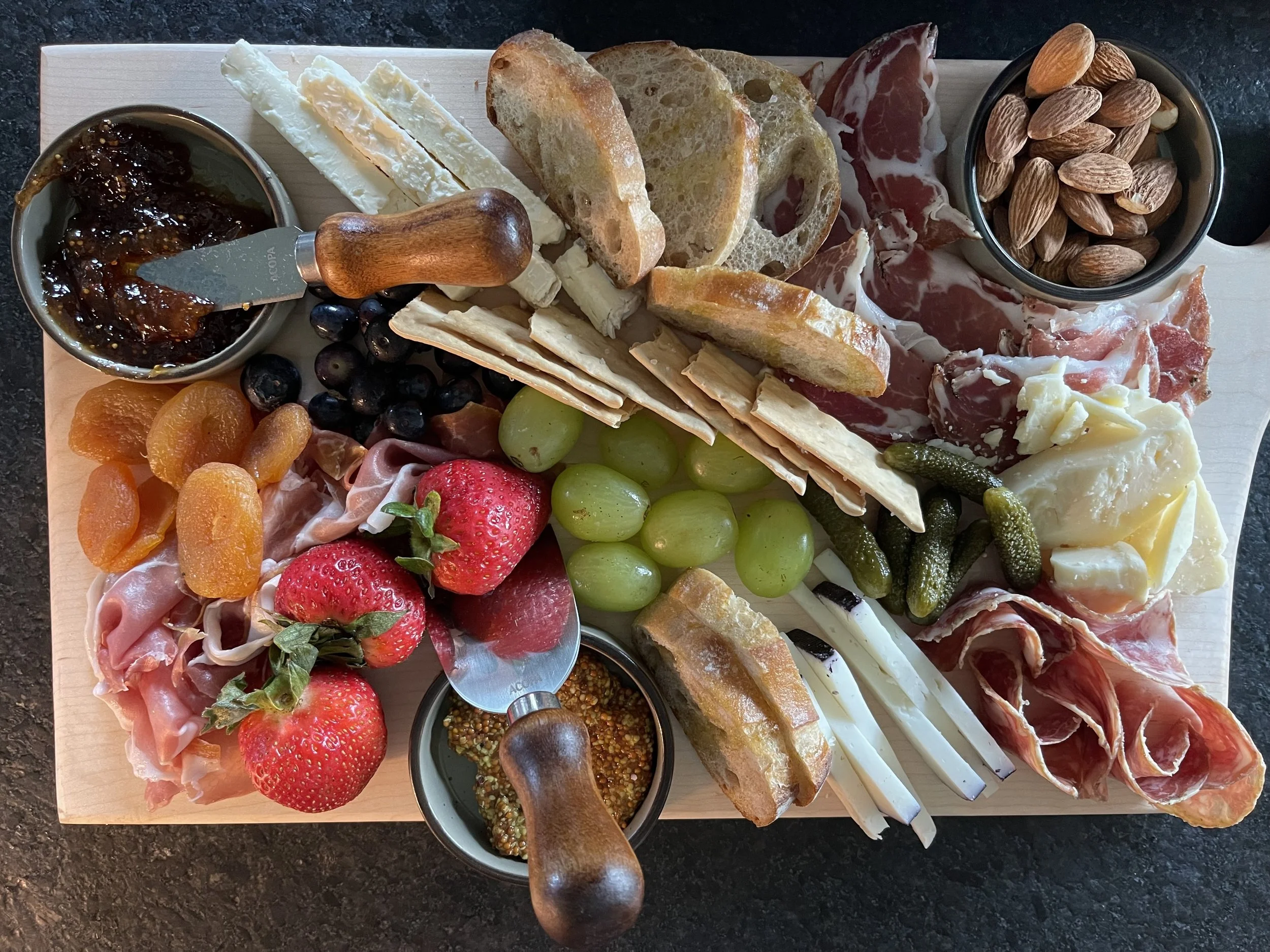 A wooden platter with assorted charcuterie foods including slices of cured meats, cheeses, bread slices, crackers, strawberries, grapes, dried apricots, almonds, and jars of jam and mustard.