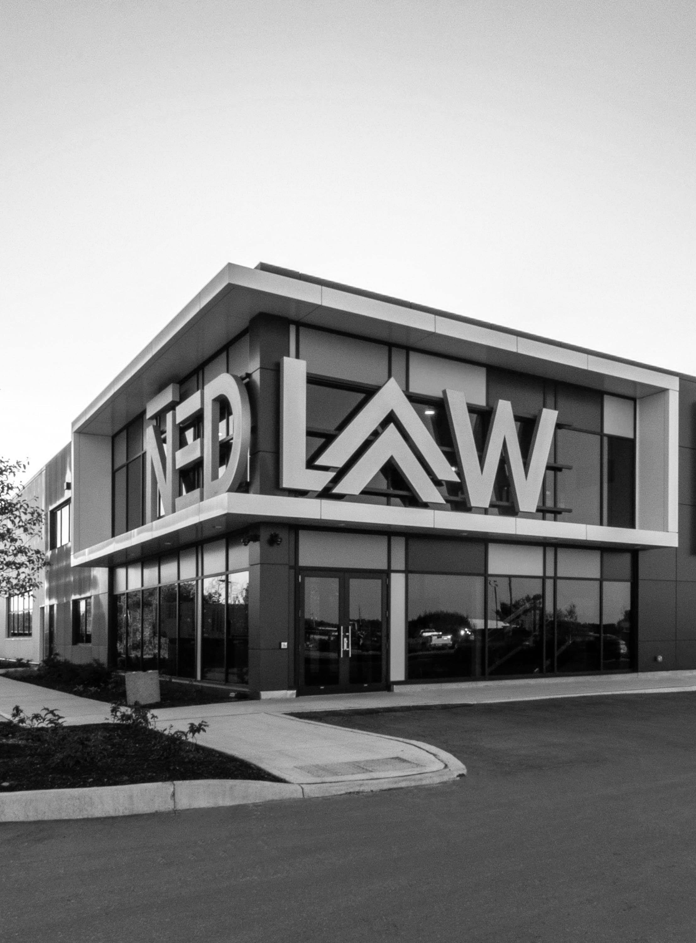 A modern commercial building with glass windows and large sign with the word 'NEWD' visible at the top corner. The parking lot and sidewalk are in the foreground, with some trees and cars reflected in the windows.