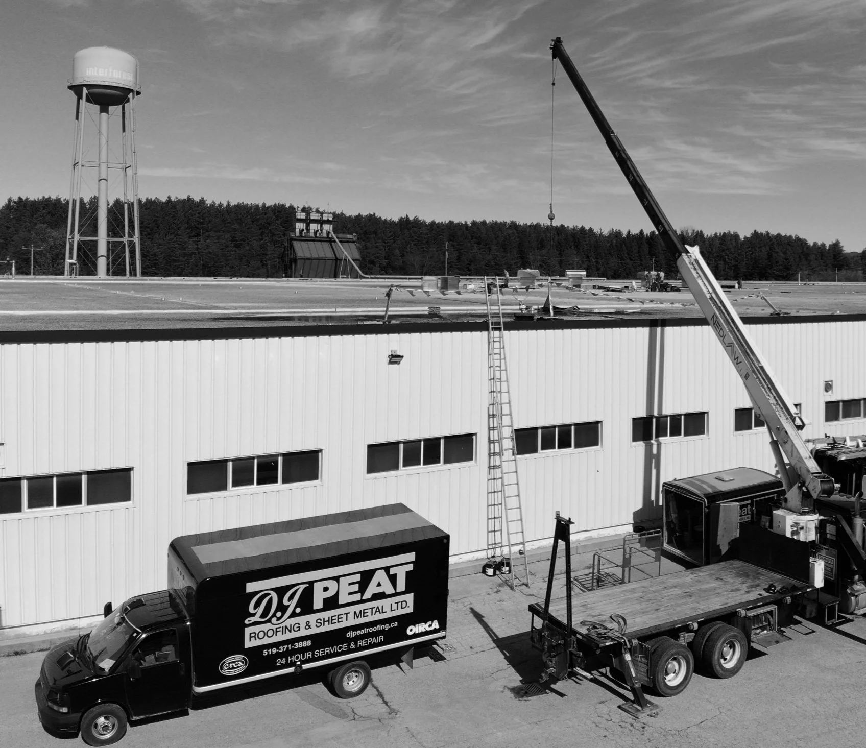 Construction site on rooftop of a commercial building with a water tower in the background, a crane, and service trucks, including a black truck with advertisement for roofing company, and metal sheets and ladders on site.