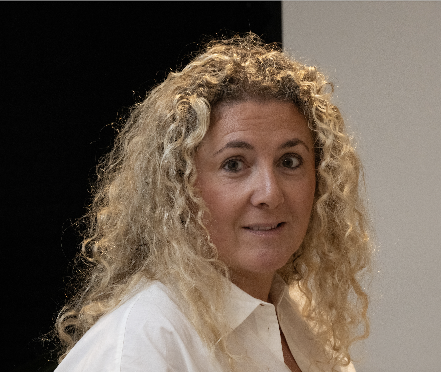 A woman with curly blonde hair wearing a white shirt, looking at the camera with a slight smile, in a well-lit indoor setting.